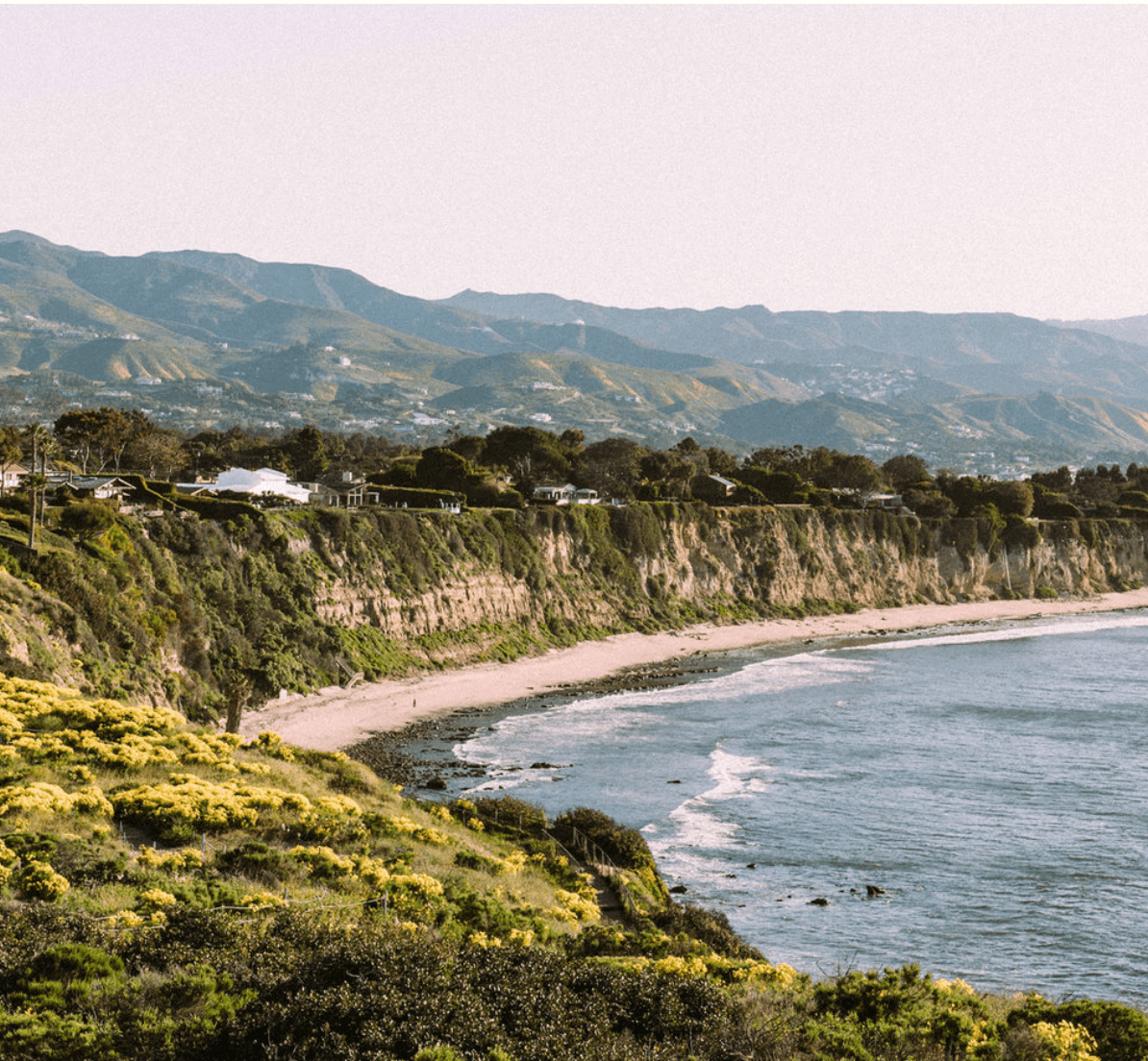 The Surfrider, North America — Coastal cliffside with sandy beach, terraced homes, verdant hillsides, and mountain range under soft natural light.