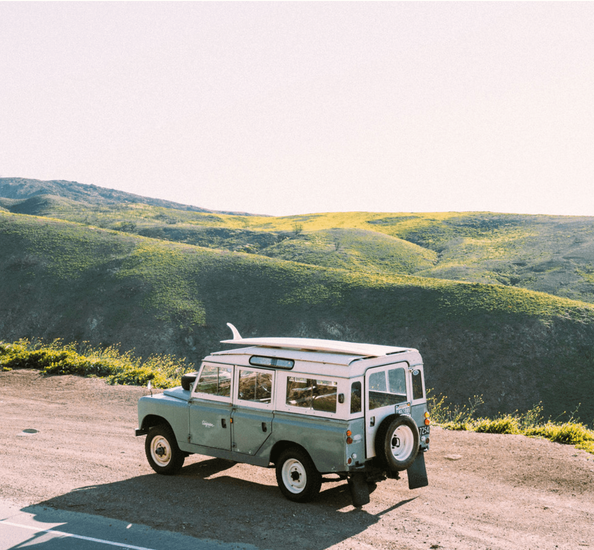 The Surfrider, North America — Vintage Land Rover parked on coastal bluff overlooking rolling golden hills, surfboard mounted on roof, soft natural light.