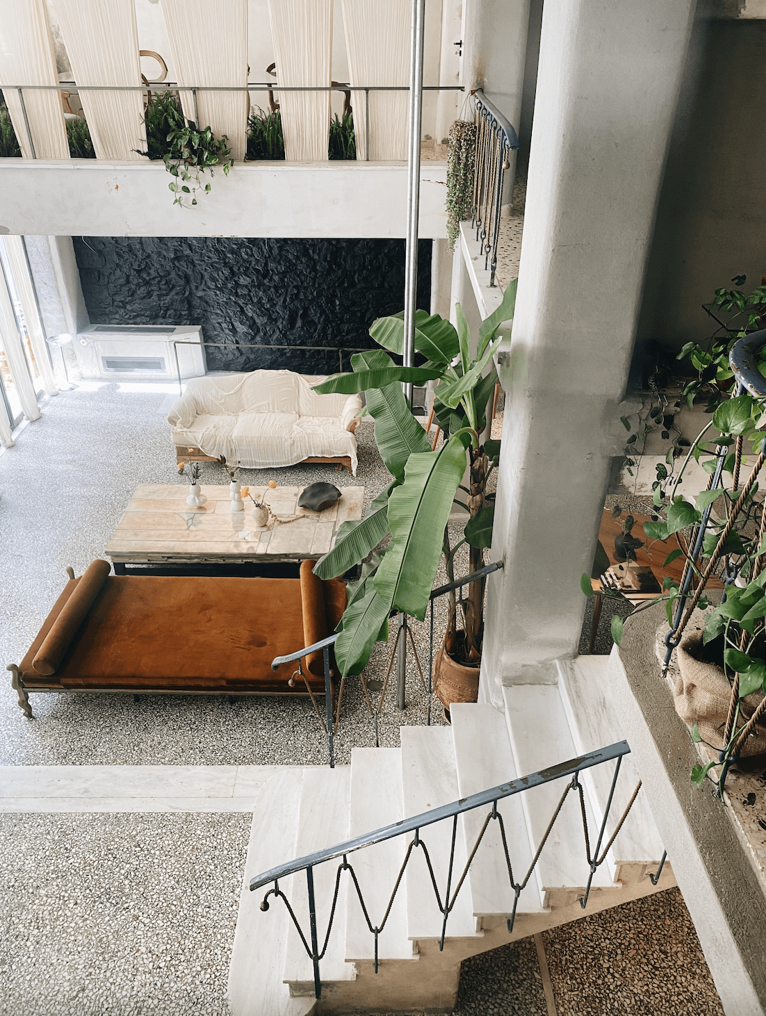 Mona, Europe — Sunlit courtyard with cream linen sofa, potted plants, wooden furniture, and wrought-iron staircase overlooking double-height loft space.