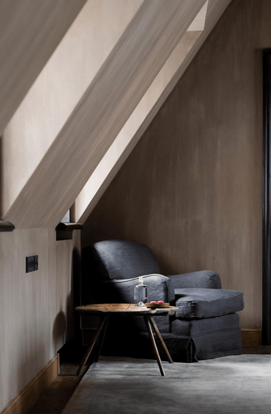 Purs, Europe — Minimalist attic nook with charcoal linen armchair, wooden tripod table, and sloped white ceiling flooded with natural light.