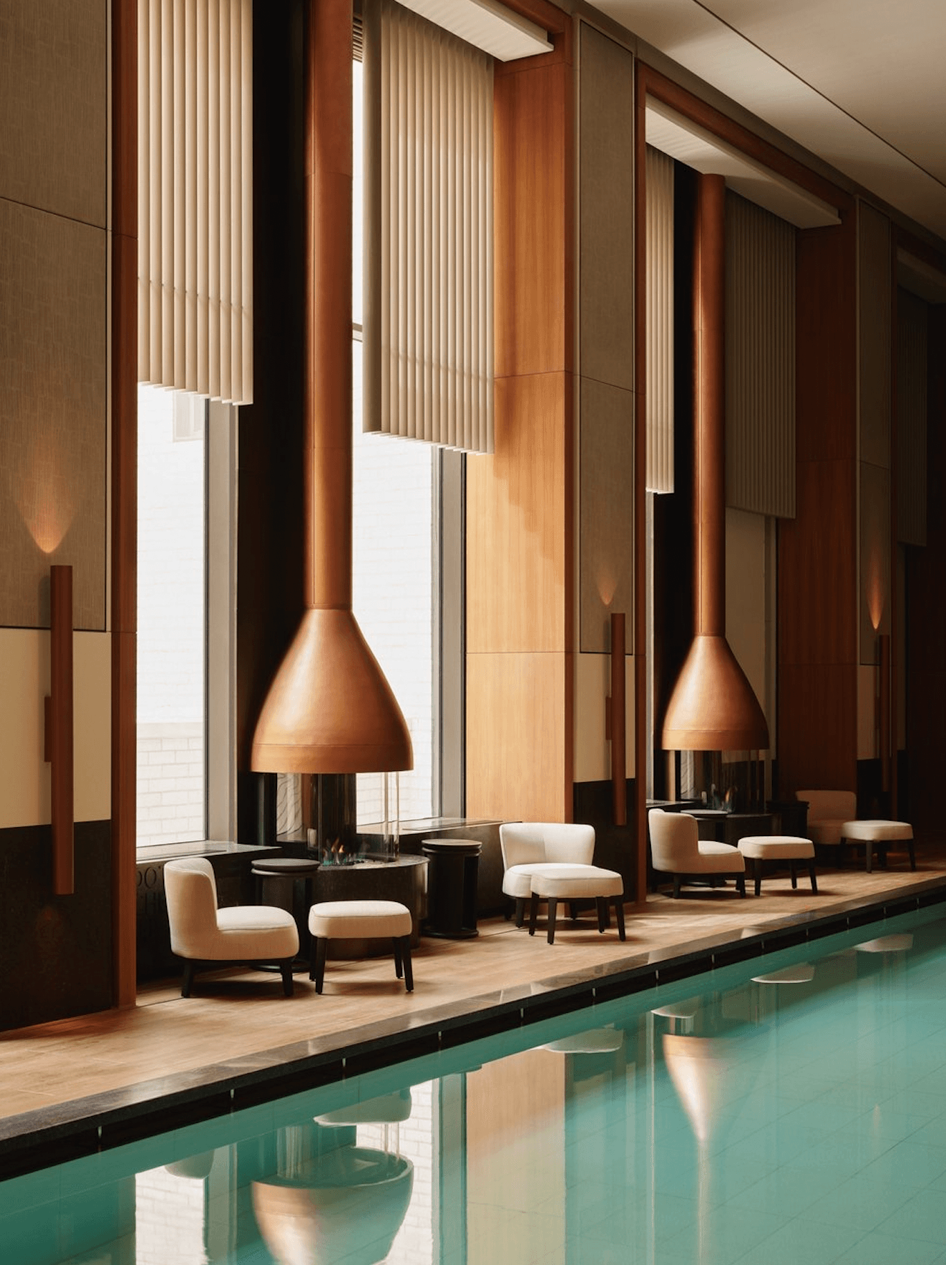 Aman New York, New York, USA — Modernist poolside lounge with copper pendant lights, cream upholstered seating, floor-to-ceiling windows, vertical wood screens, and turquoise water reflecting warm wood tones.