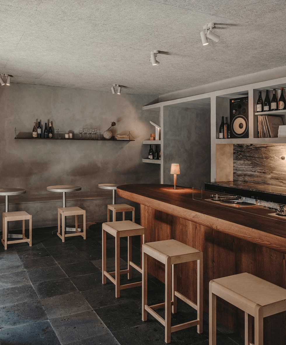 Gota Wine Bar, Europe — Concrete walls, wooden bar counter, natural linen bar stools, shelving with wine bottles, warm ambient lighting, minimalist hospitality interior design.
