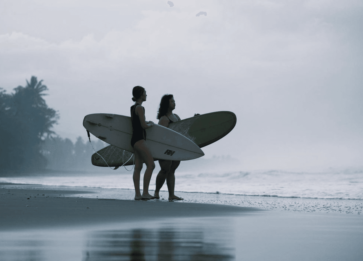 Two surfers in wetsuits stand on the wet sand at Lost Beach, surfboards in hand, beneath overcast skies.