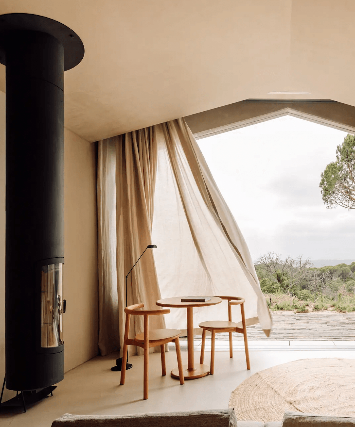 Pa.te.os, Europe — Minimalist bedroom with cylindrical black wood-burning stove, natural linen furnishings, and angled window framing savanna landscape.