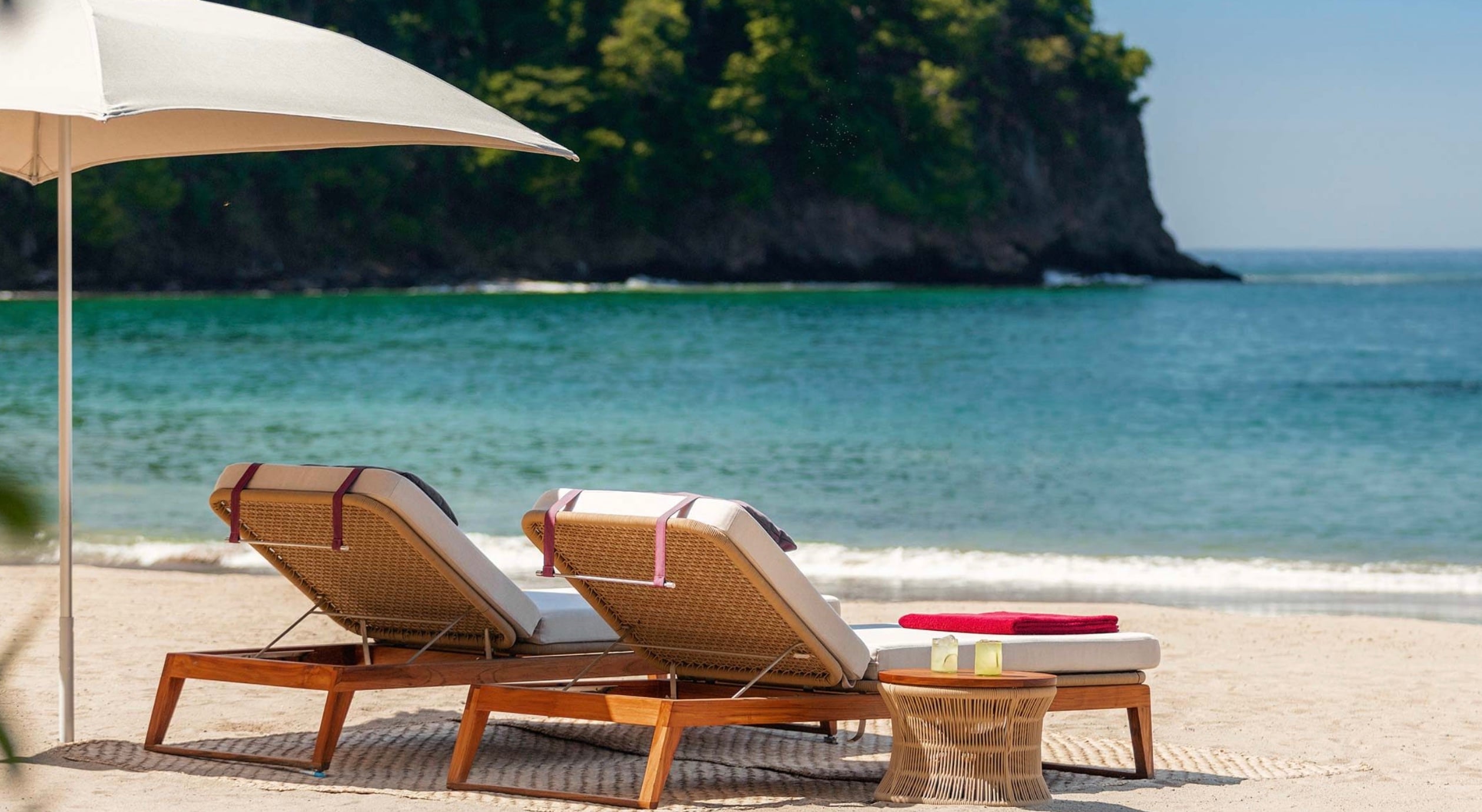 Jetty Beach Club, Mexico — Two woven lounge chairs with white cushions beneath a sail shade overlooking turquoise waters and limestone cliffs.