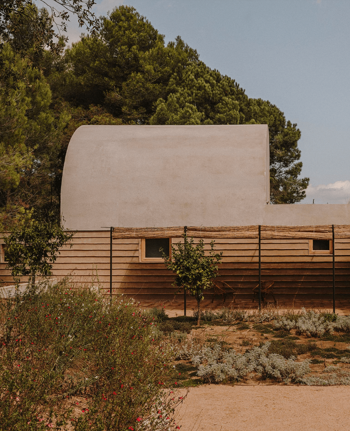 Casa Ter | MESURA — Casa Ter — Curved concrete volume with wooden horizontal siding, metal railing, and native Mediterranean plantings in arid landscape with pine forest backdrop.