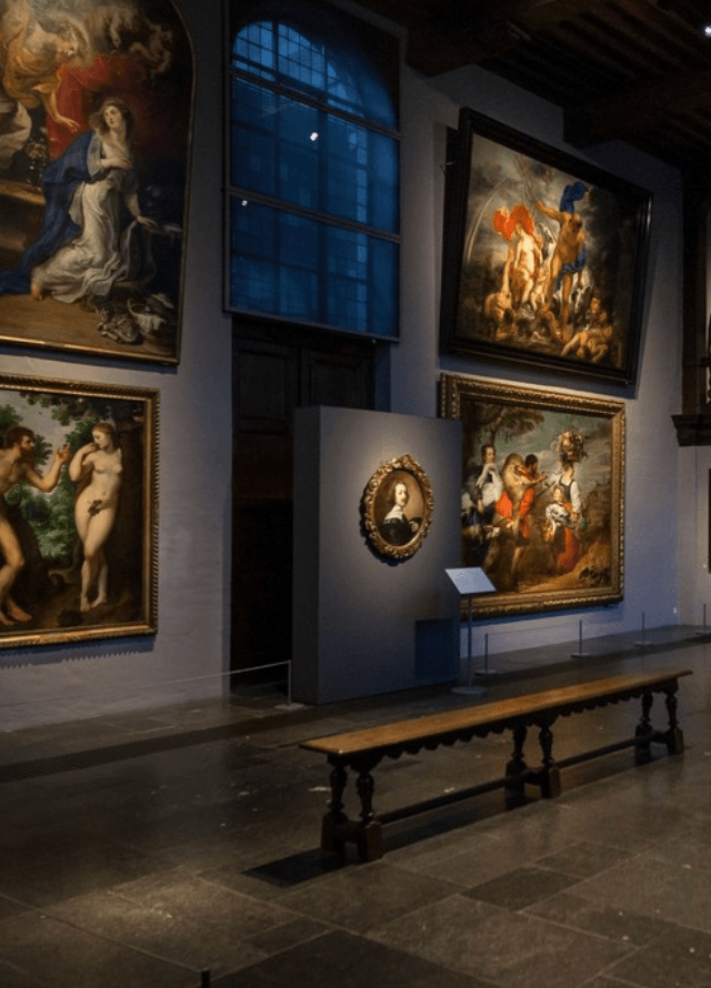 Rubens House, Europe — Renaissance paintings in gilt frames displayed on gallery walls beside arched window, wooden bench in foreground, warm museum lighting.