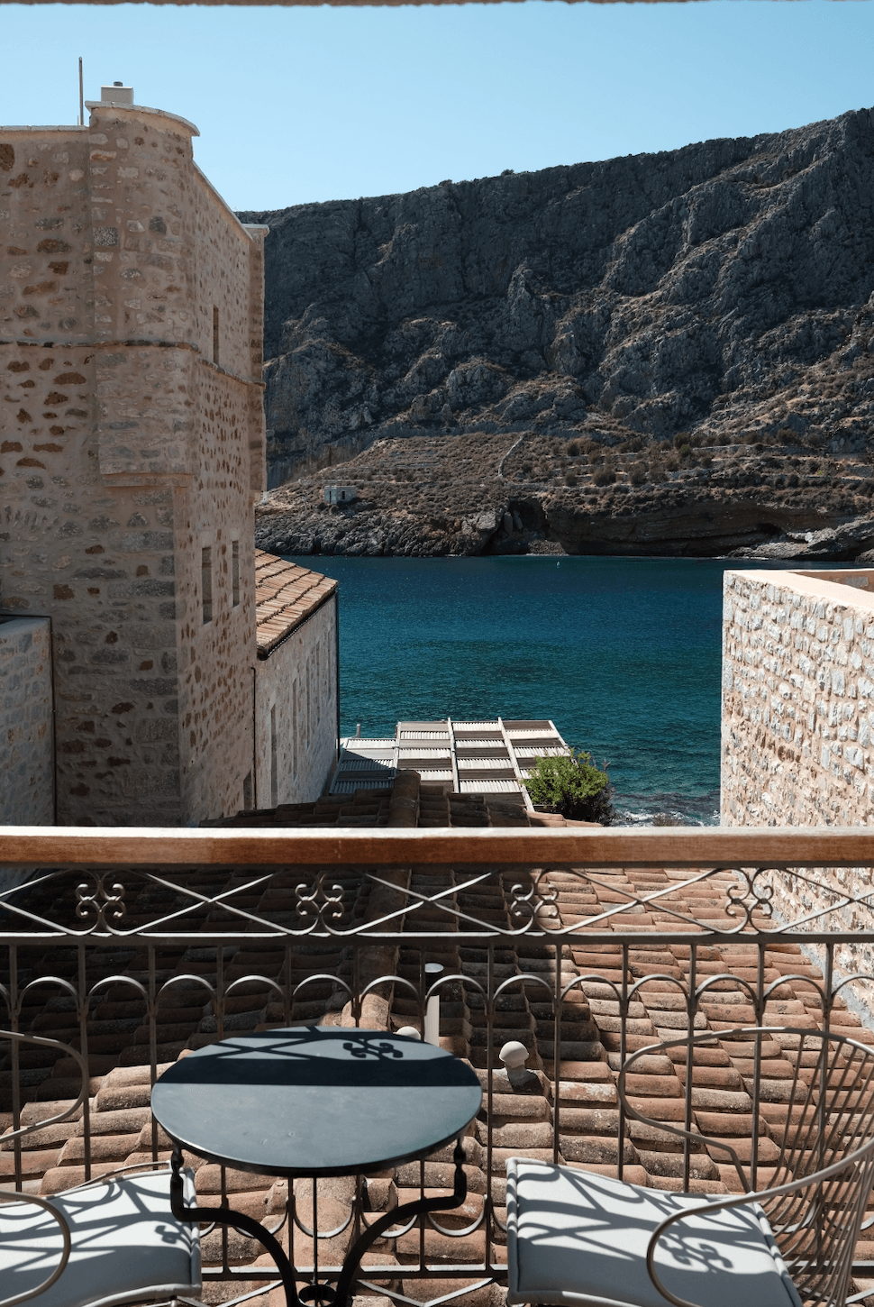 Kyrimai Hotel Restaurant, Greece — Coastal terrace with stone architecture, turquoise Aegean water, dramatic limestone cliffs, iron railing, afternoon light casting geometric shadows.