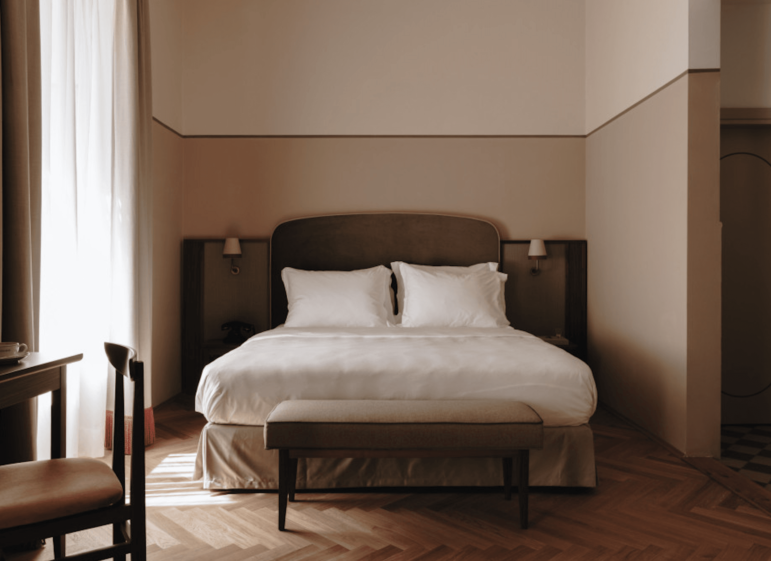Parkhotel Mondschein, Europe — Minimalist hotel bedroom with dark headboard, white linens, herringbone wood floors, and soft natural light from tall windows casting geometric shadows.