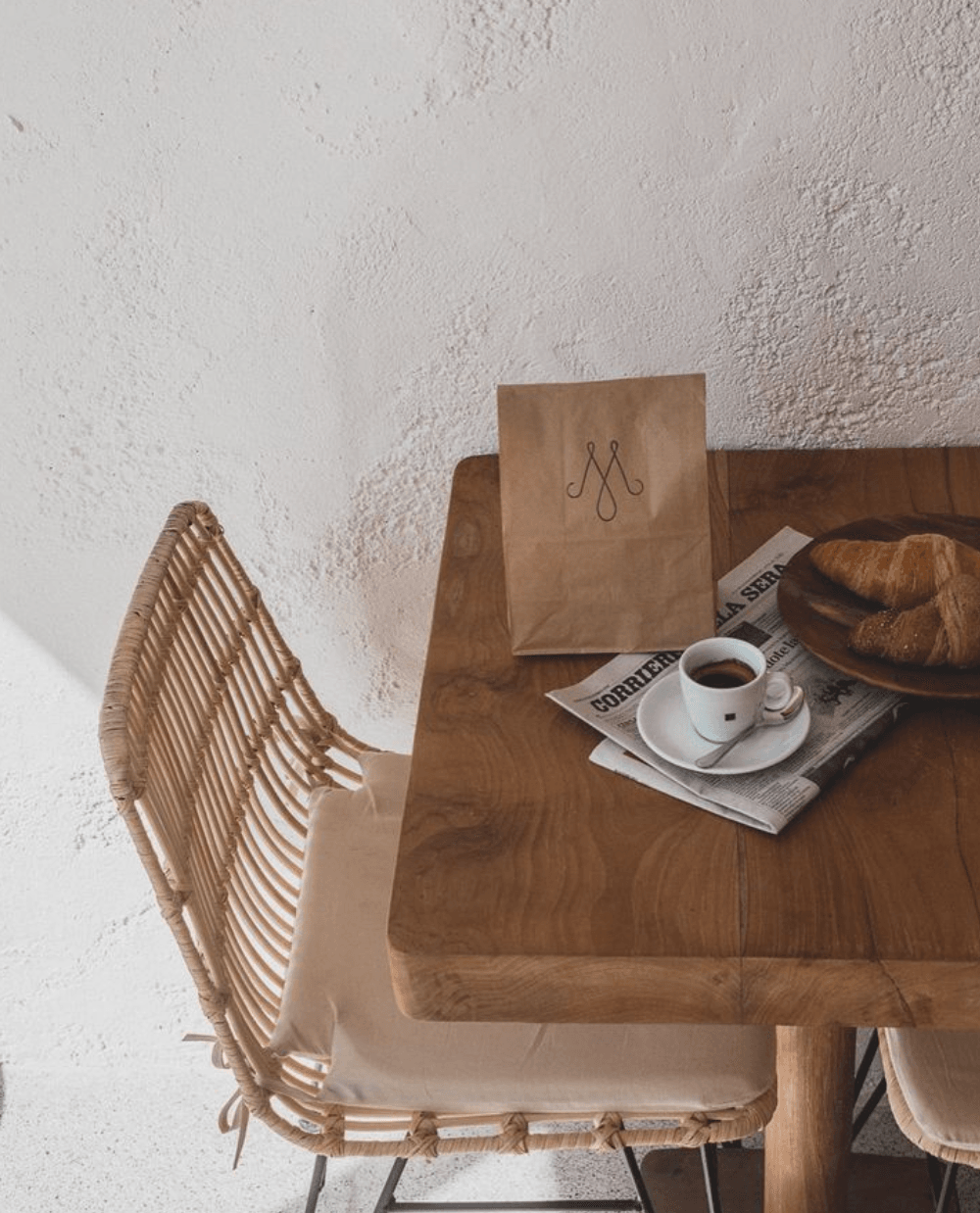 Massa Coffee, Balearic Islands — Woven rattan chair at wooden table with espresso, croissant, newspaper, and kraft paper bag in minimalist café setting.