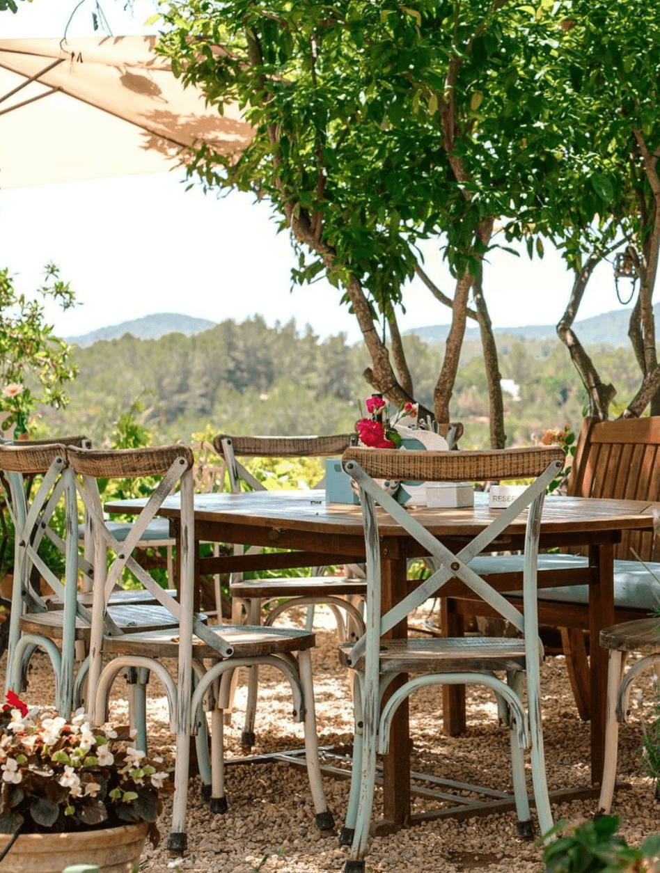 La Paloma, Balearic Islands — Whitewashed bistro chairs at wooden table beneath olive trees with mountain views and terracotta courtyard.