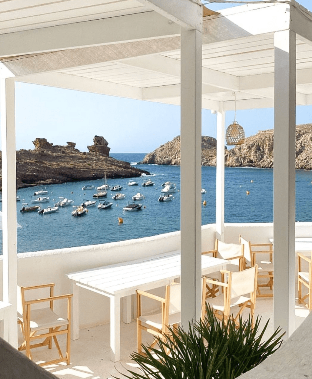 Ivette, Balearic Islands, Spain — Whitewashed pergola dining pavilion overlooking moored boats in turquoise cove with limestone cliffs, natural light and minimalist wood furniture.