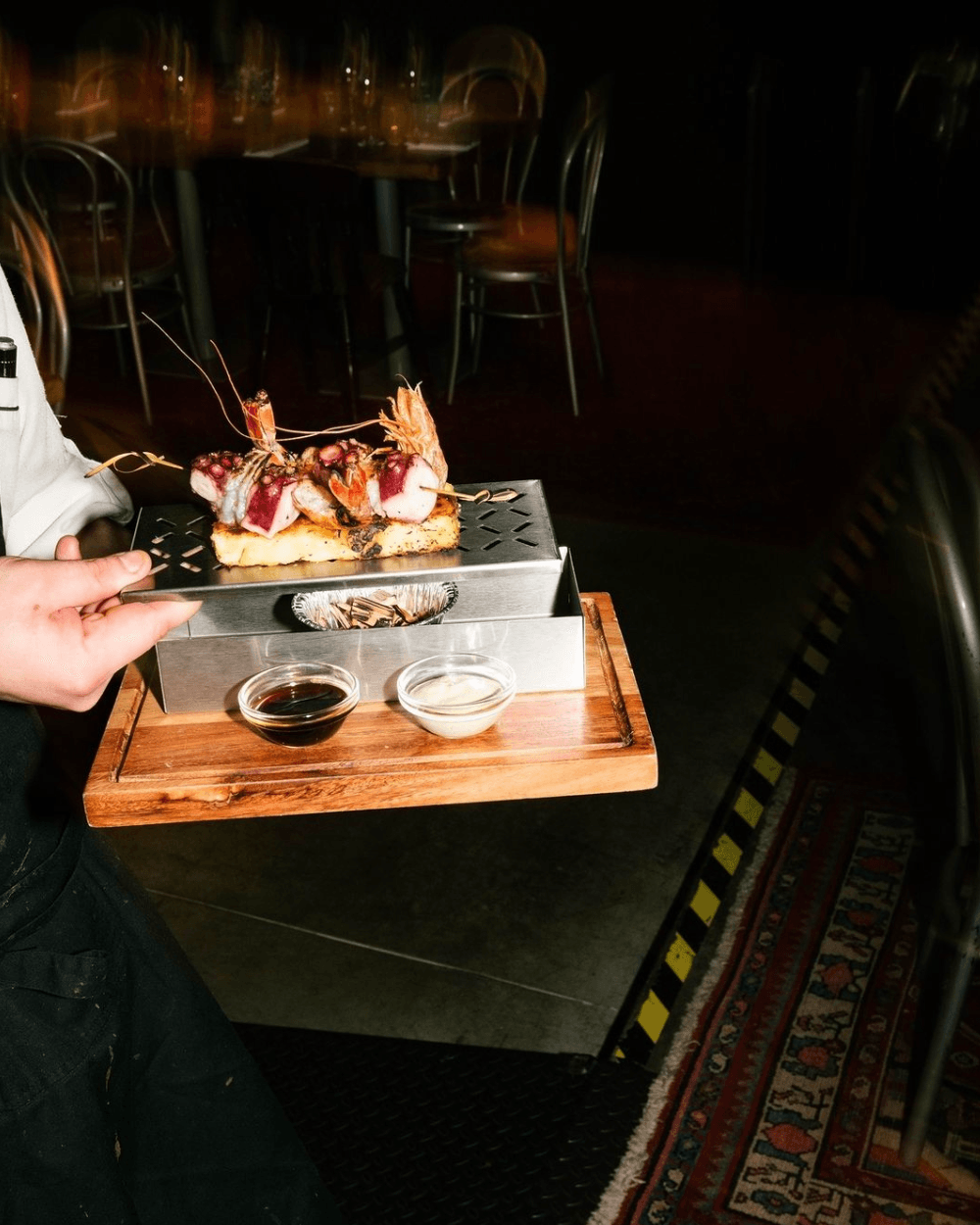 Laboratorio Lanzani, Italy — Seared meat and seafood skewer on toasted bread with sauce accompaniments, plated on stainless steel and wood in intimate evening setting.