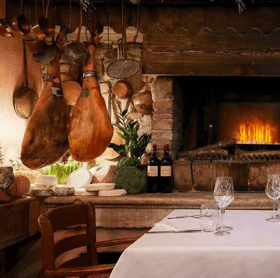 Trattoria Porteri, Italy — Rustic dining room with cured meats hanging from beam, fireplace glow, stone architecture, wine bottles, fresh vegetables and white tablecloth.