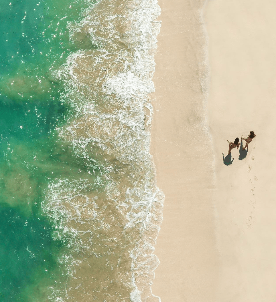 Innit Lombok, Asia — Aerial view of two guests walking along pristine sandy beach meeting turquoise ocean waves at waterline.