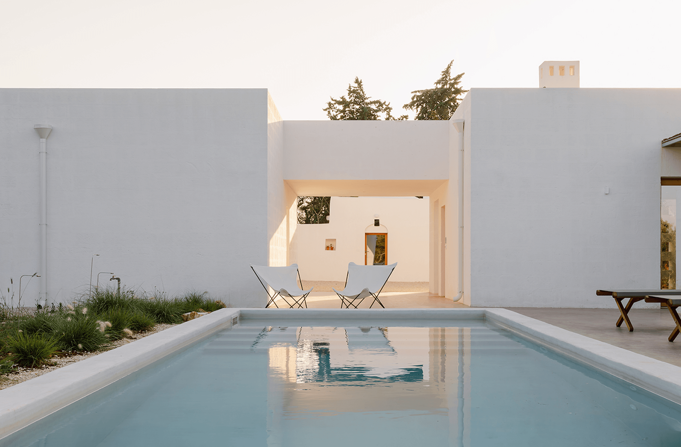 Casanguilla, Europe — Minimalist white villa courtyard with rectangular pool, canvas chairs, and modernist architecture framing serene Mediterranean landscape.