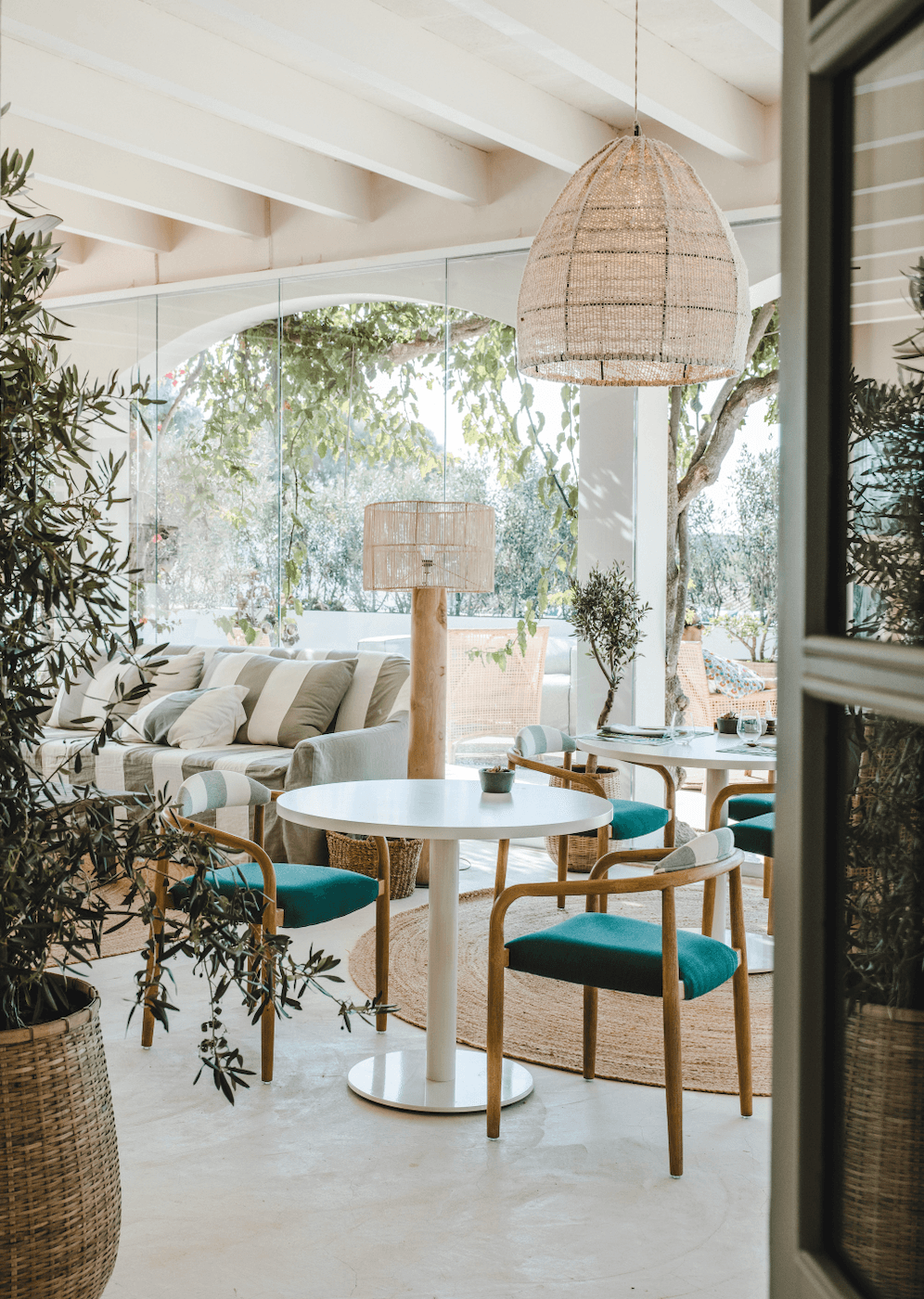 Morvedra Nou Table, Balearic Islands, Spain — Covered terrace with woven pendant light, white round table, wooden chairs with teal cushions, potted olive trees, glass railings, and coastal views.