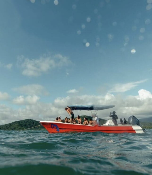 Private Boat La Gozadera, Mexico — Red speedboat with canopy anchored in turquoise water, passengers aboard, forested coastline and white clouds under blue sky.