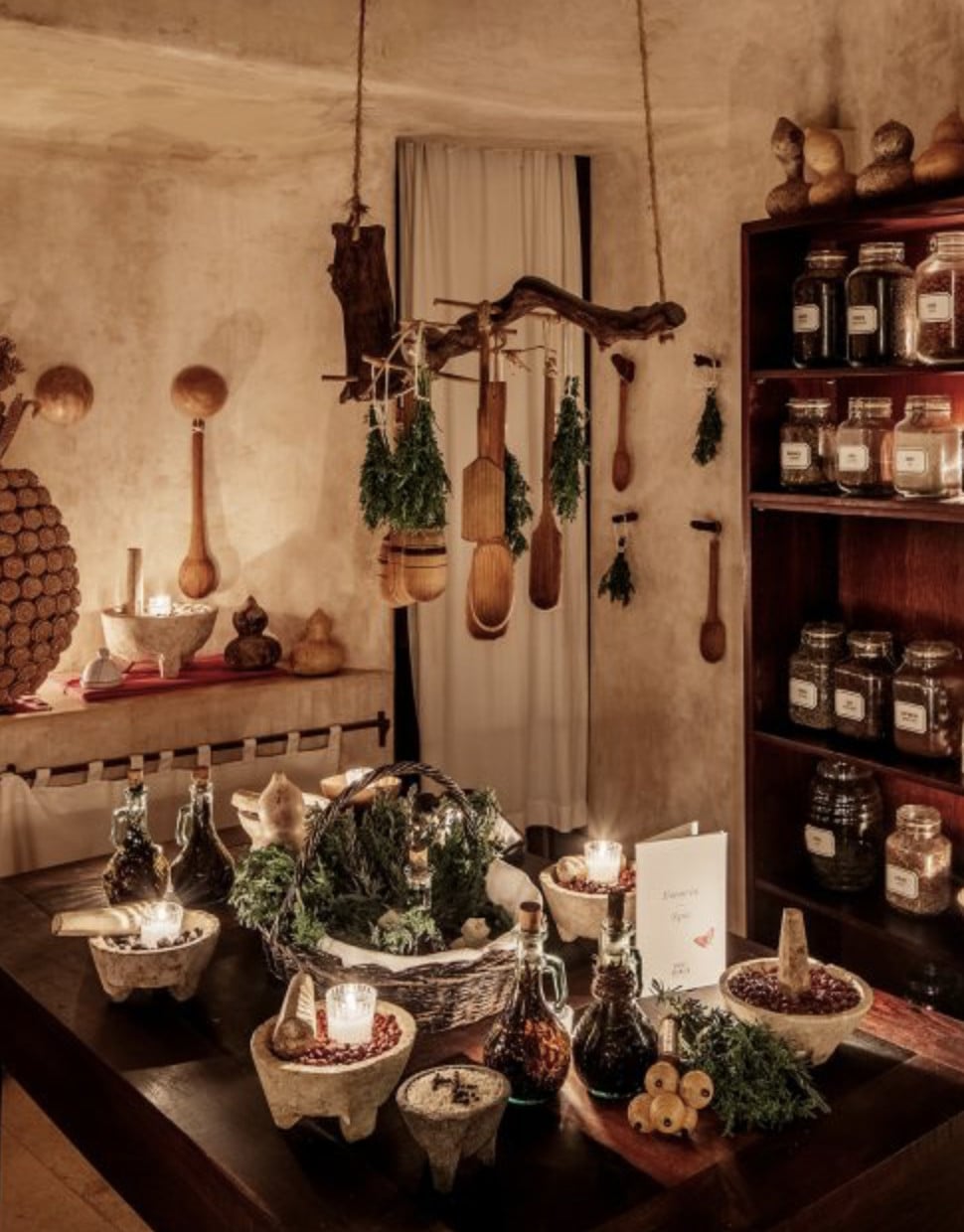 Esencia Spa, Mexico — Apothecary shelving with glass jars, hanging wooden utensils and dried herbs, potted plants with candles, earthy wellness interior design.
