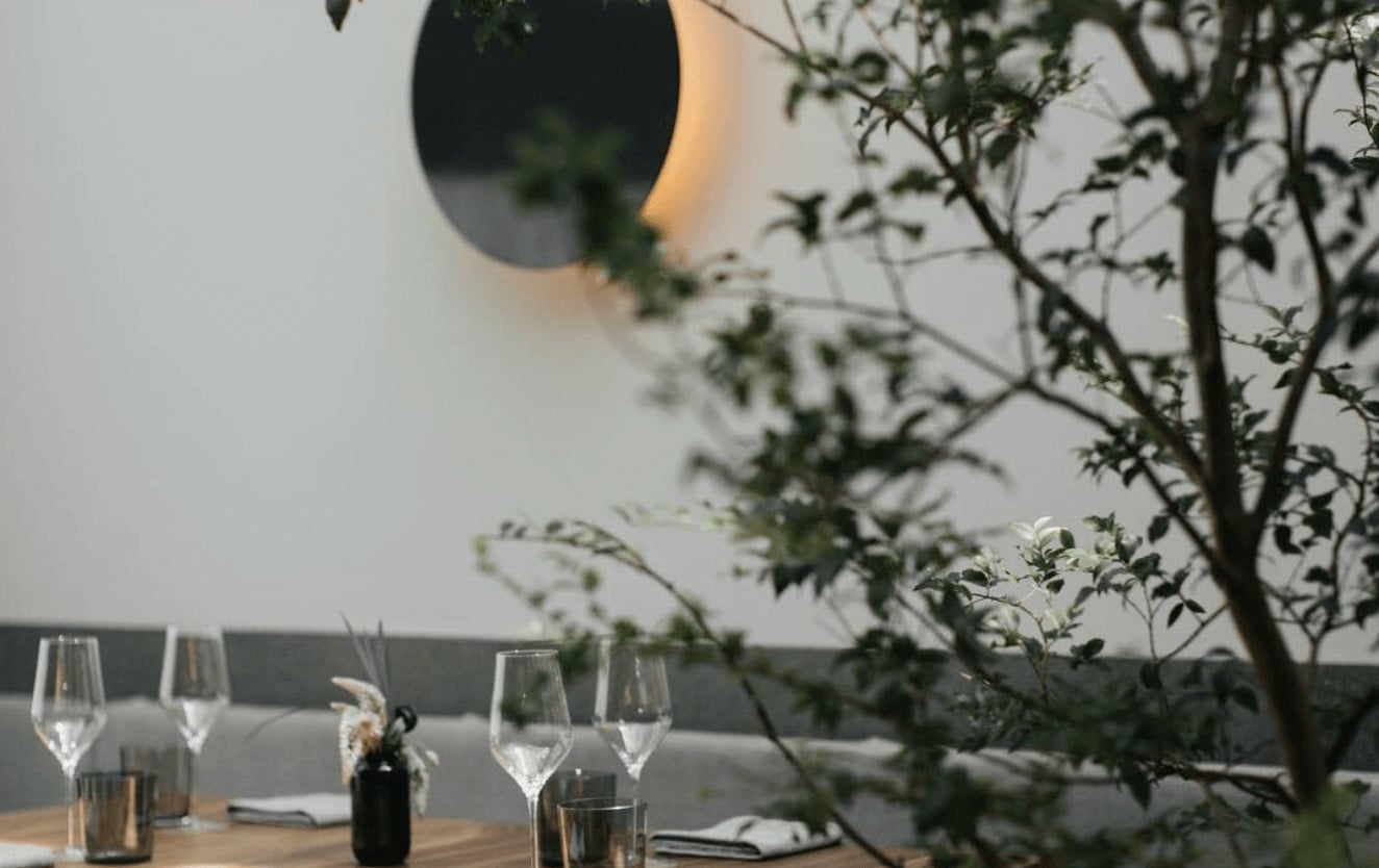 Pujol, Mexico City, Mexico — Minimalist dining room with green foliage, wine glasses, and warm circular wall light above grey banquette seating.