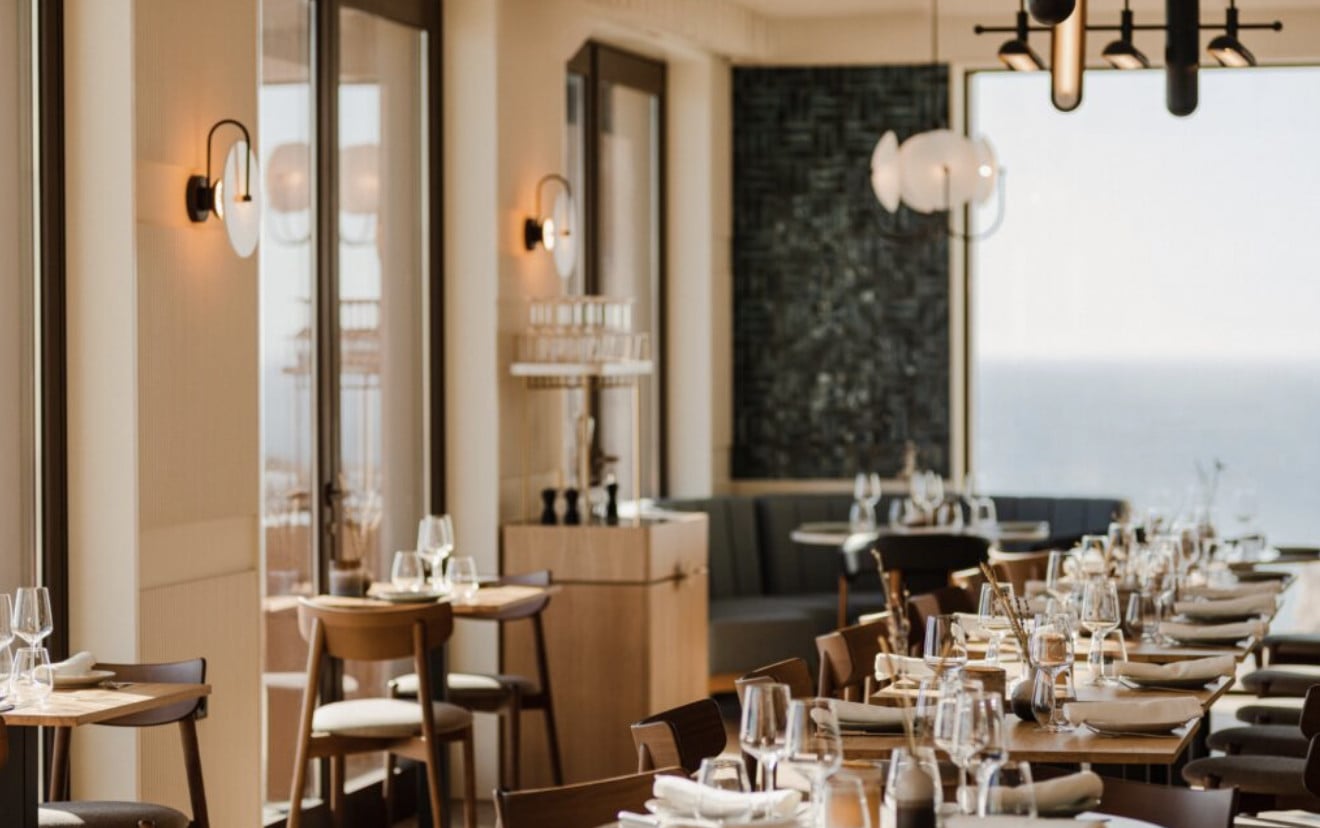 Onda, Europe — Minimalist coastal restaurant with floor-to-ceiling windows, wood furnishings, marble accents, and pendant lighting overlooking seascape.