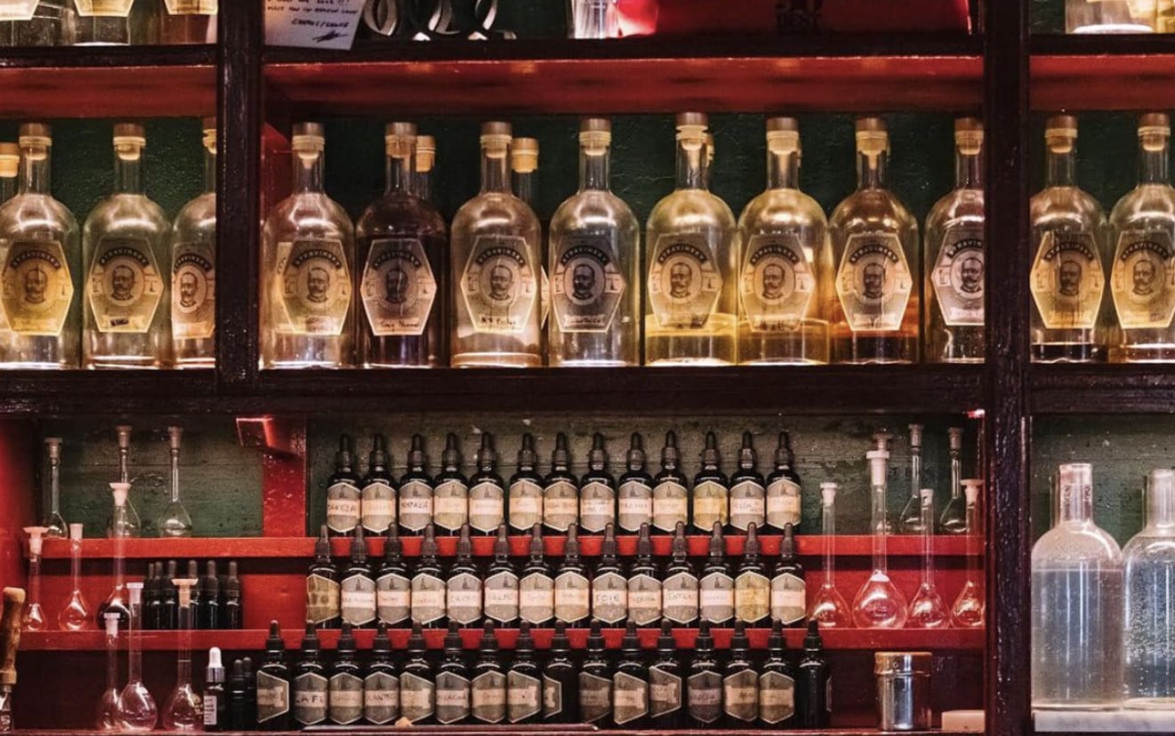 Dr. Stravinsky, Europe — Illuminated bar display with vintage spirit bottles in glass shelving against dark wood and deep green wall paneling, warm ambient lighting.