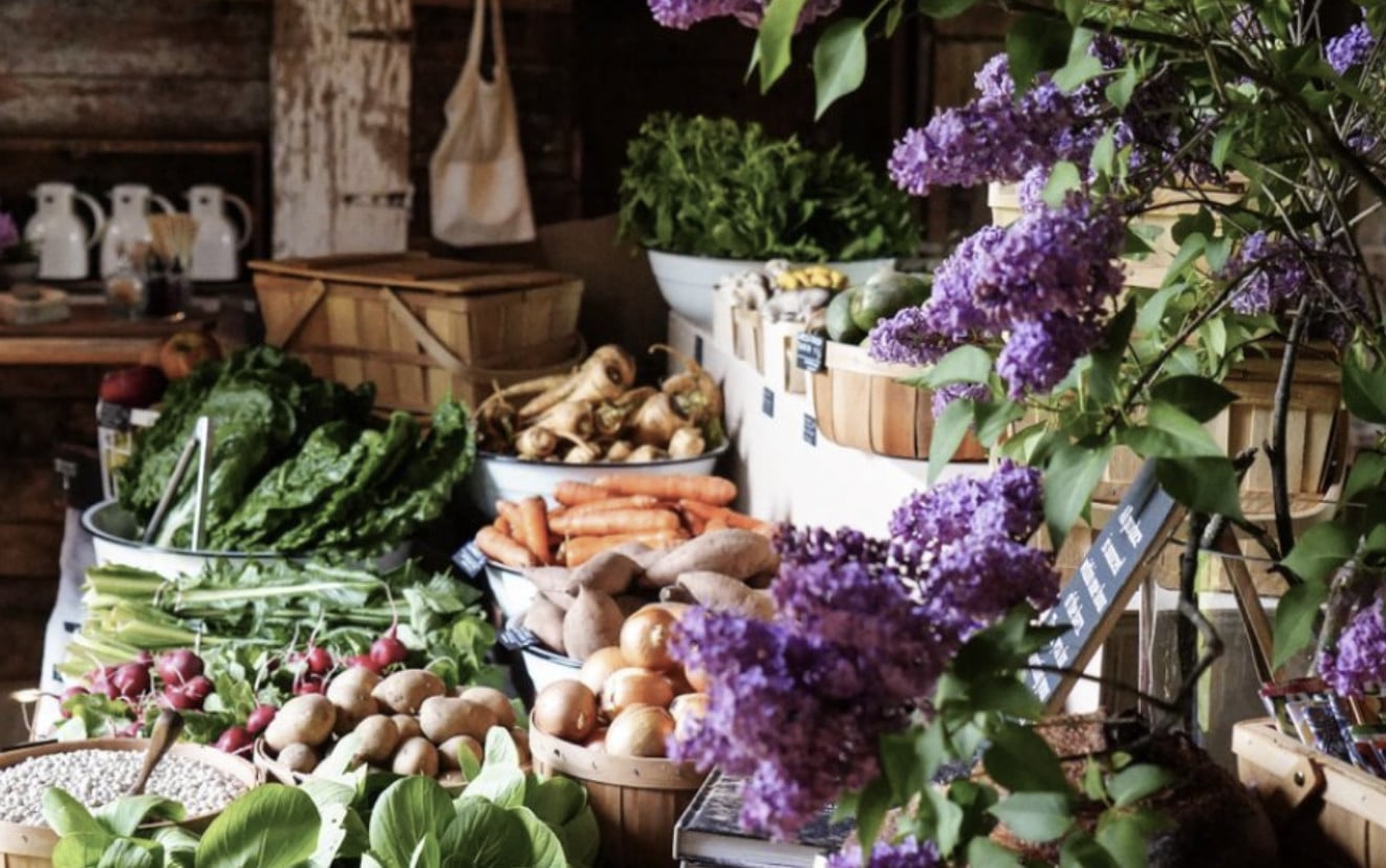 Ravenwood Farm, North America — Farm stand with purple lilac flowers, fresh vegetables in wooden baskets, radishes, carrots, and potatoes displayed outdoors.