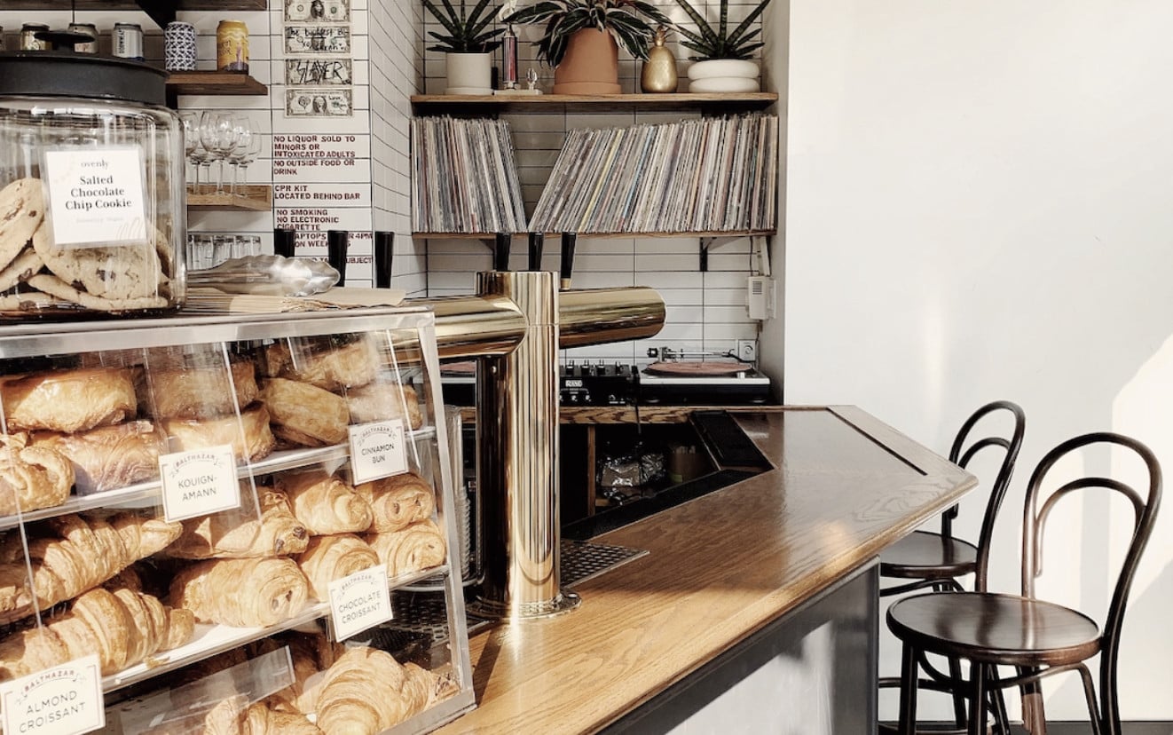 Little Canal, North America — Minimalist bakery counter with croissants in glass display, wooden service bar, vinyl records on shelves, and black bentwood chairs.