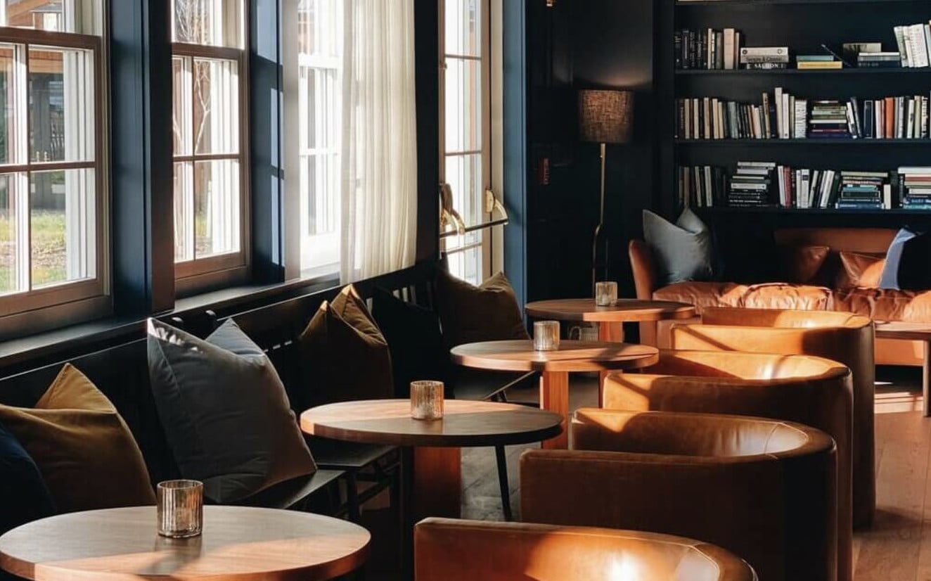 Library Bar, North America — Sunlit lounge with cognac leather chairs, dark shelving, wooden tables, and floor-to-ceiling windows in a sophisticated hospitality space.