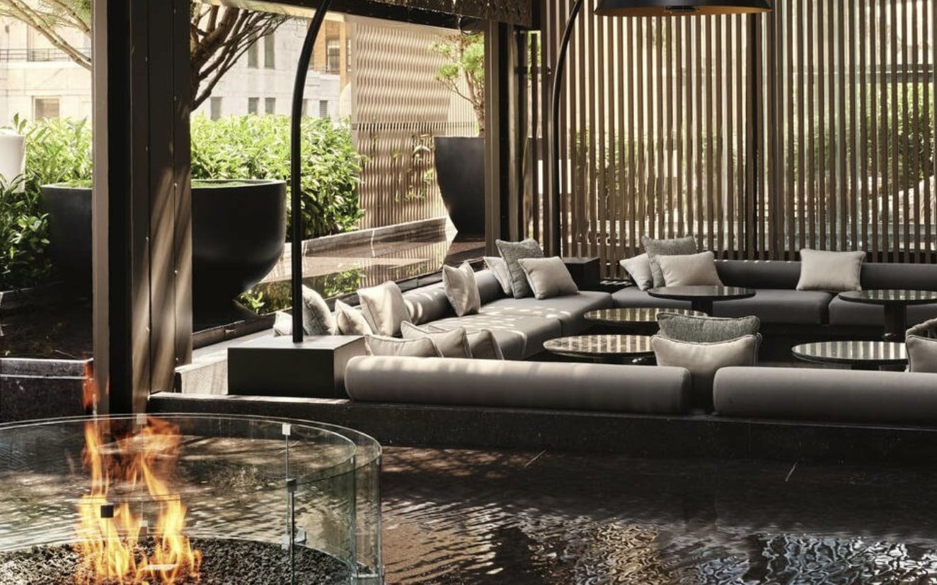 The Garden Terrace, North America — Modern lounge with curved gray sectional, glass fire pit, black vertical screens, potted greenery, and afternoon shadow patterns on stone patio.