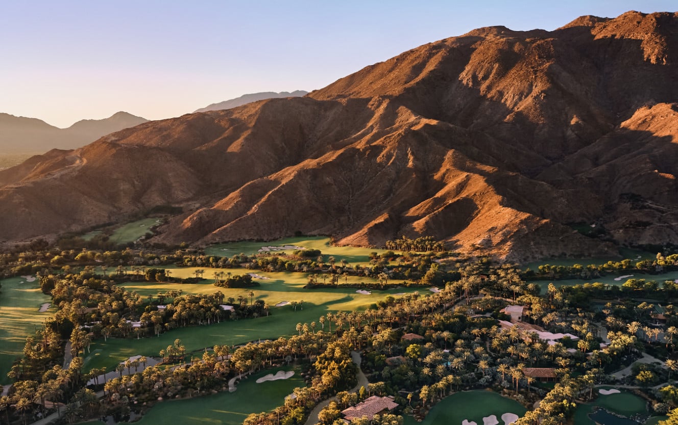 Aerial view of a championship golf course nestled in a desert valley at golden hour, surrounded by palm trees and dramatic canyon walls.