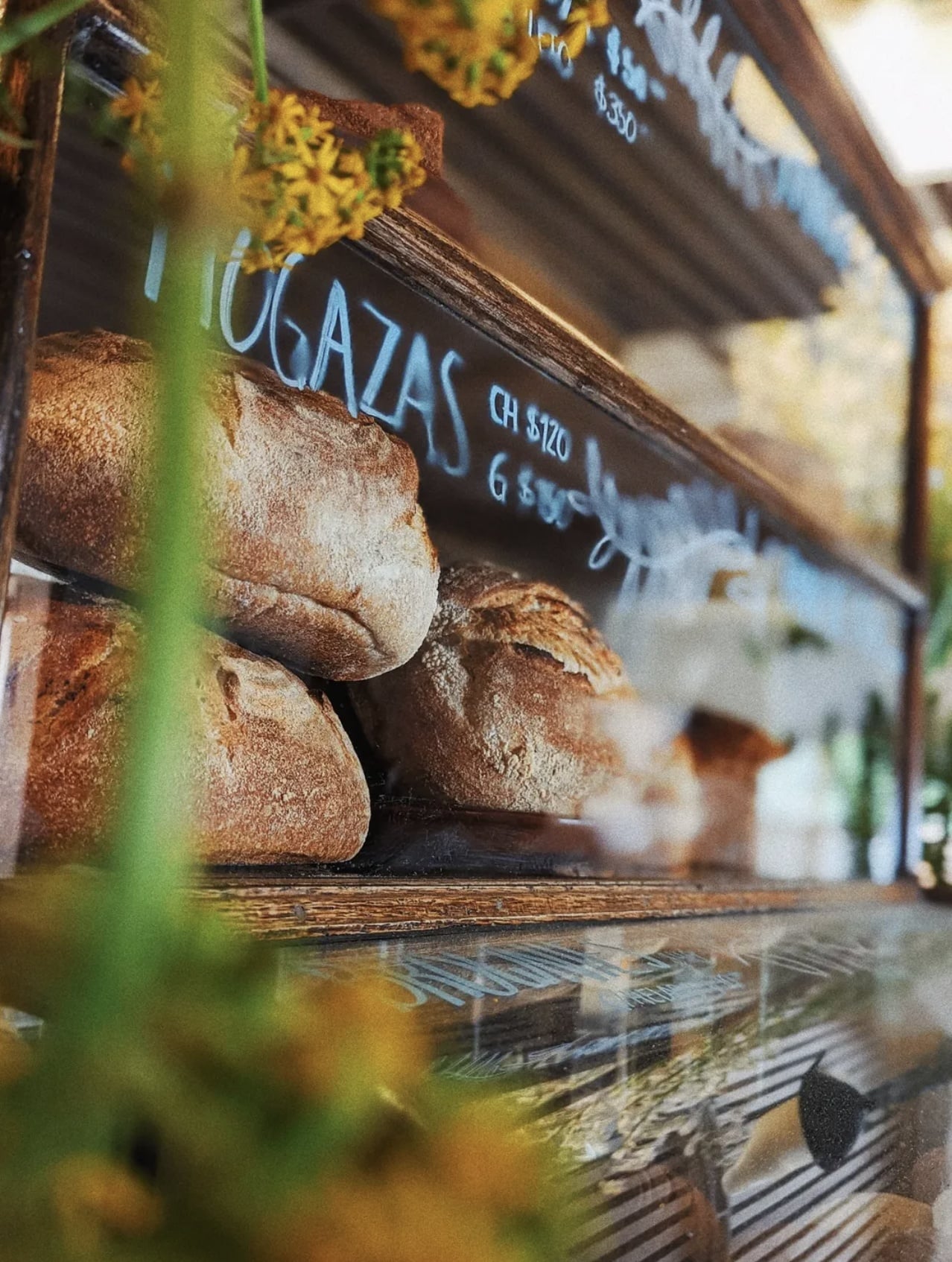 Faena Local, Mexico — Artisanal bread loaves with flour dusting displayed in wooden bakery case with yellow flowers and handwritten pricing labels.