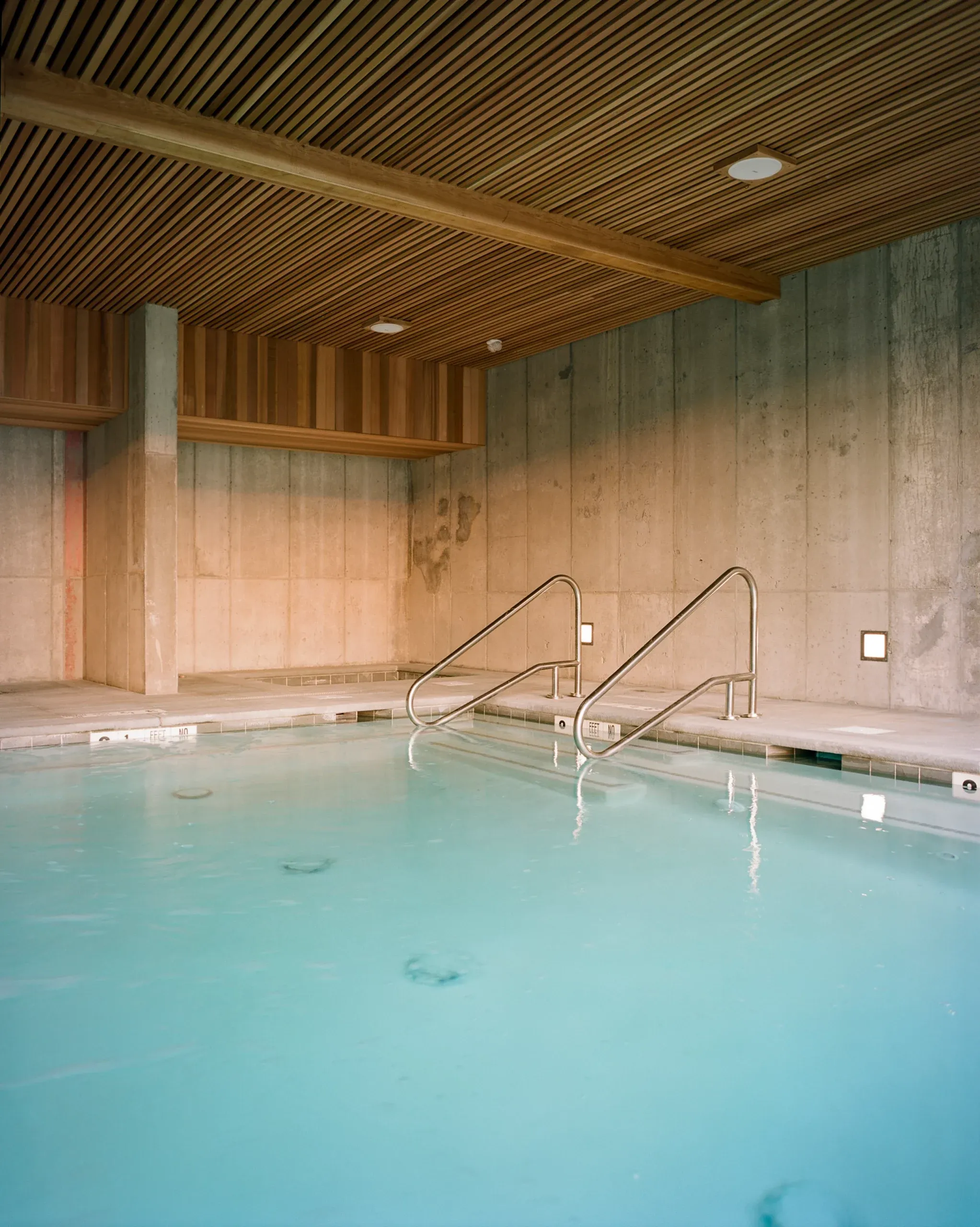 Piaule, North America — Indoor swimming pool with turquoise water, stainless steel handrails, concrete walls, and slatted wood ceiling in modernist hospitality design.