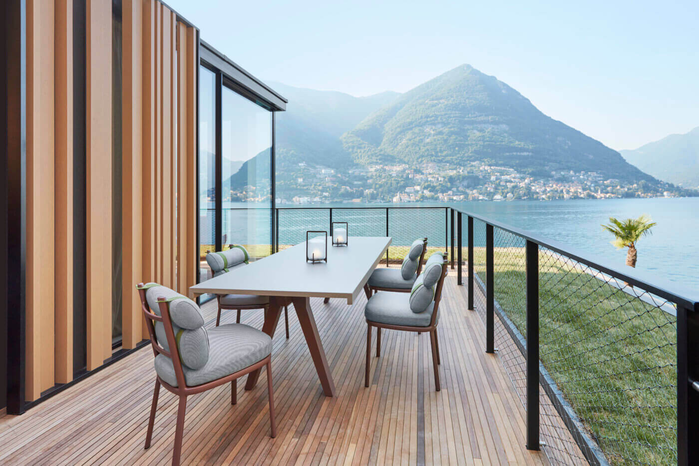 il Sereno Lago di Como, Italy — Modern lakeside terrace with wooden decking, contemporary dining table, grey upholstered chairs, mountain views across alpine lake, metal cable railing, vertical wood cladding.