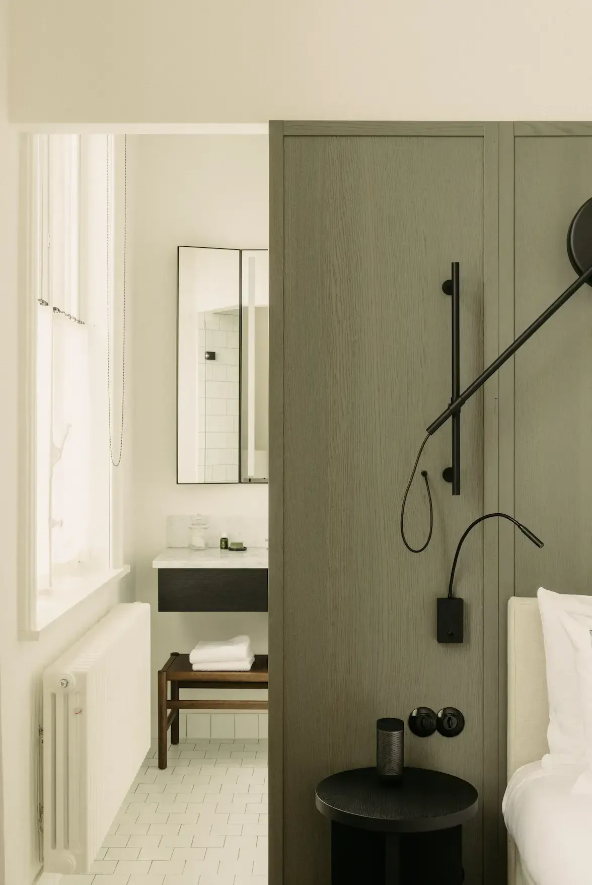 August Hotel, Europe — Minimalist bathroom with sage green paneling, black fixtures, white subway tile floor, and sculptural black vessel sink beside bed alcove.