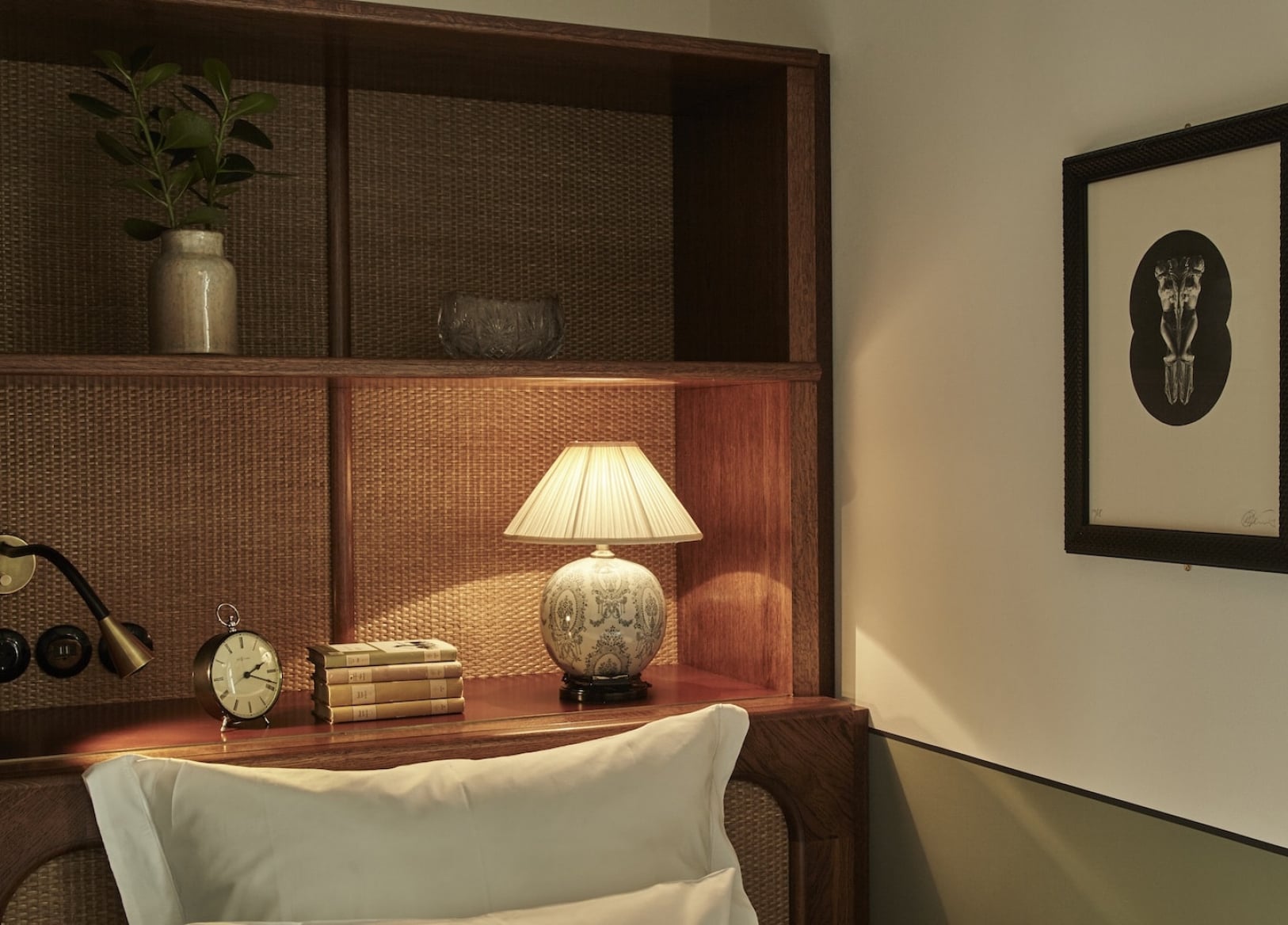Hotel Sanders, Copenhagen, Denmark — Warm wooden shelving unit with pleated lamp, stacked books, vintage clock, and ceramic vessels in minimalist hotel room.