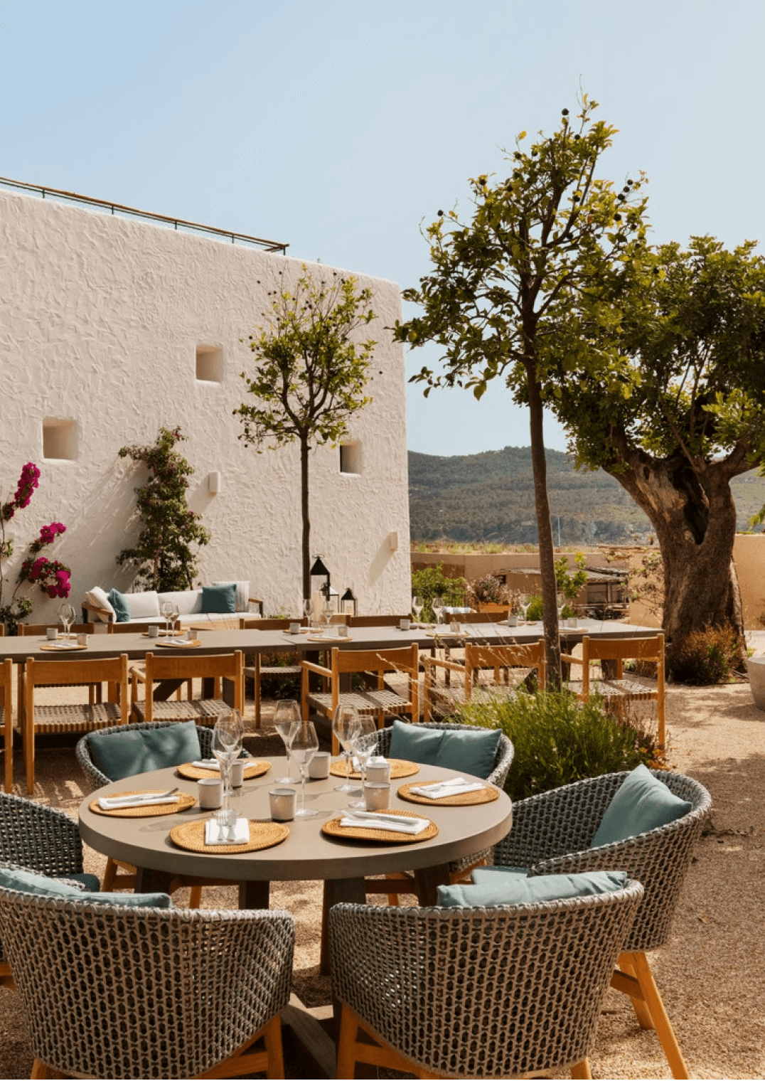 Six Senses Ibiza, Balearic Islands, Spain — Whitewashed courtyard with woven dining chairs, round stone table, gnarled olive tree, and minimalist architecture overlooking terracotta landscape.