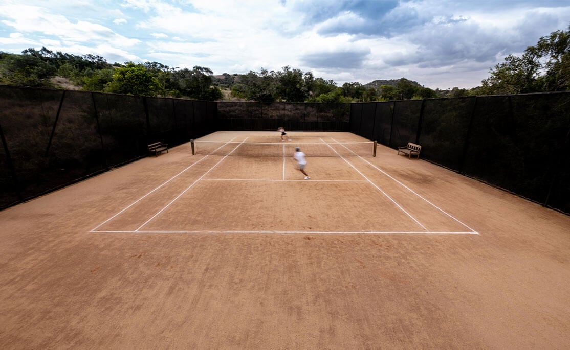 Arijiju Retreat, Africa — Clay tennis court with white court lines, black wind screens, and savanna landscape under clouded sky.