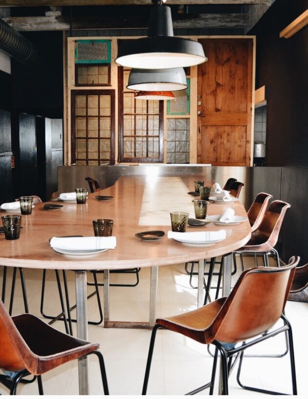 Smoix, Balearic Islands — Modern dining room with natural wood oval table, cognac leather chairs, industrial pendant lights, and salvaged architectural details on black wall.