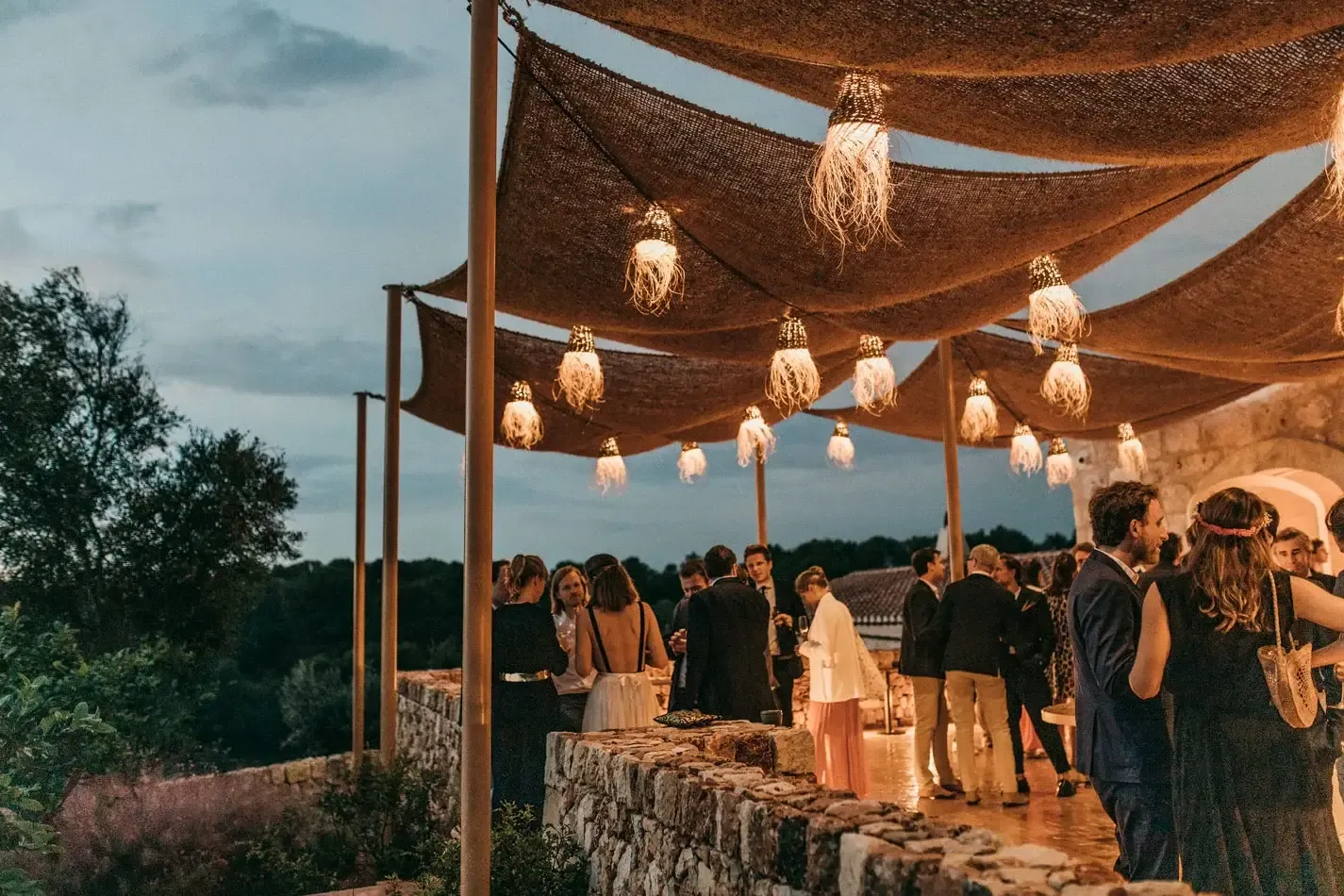 Son Blanc Farmhouse, Balearic Islands, Spain — Twilight terrace with draped fabric canopy, woven pendant lights, stone walls, and guests in evening wear beneath open sky.