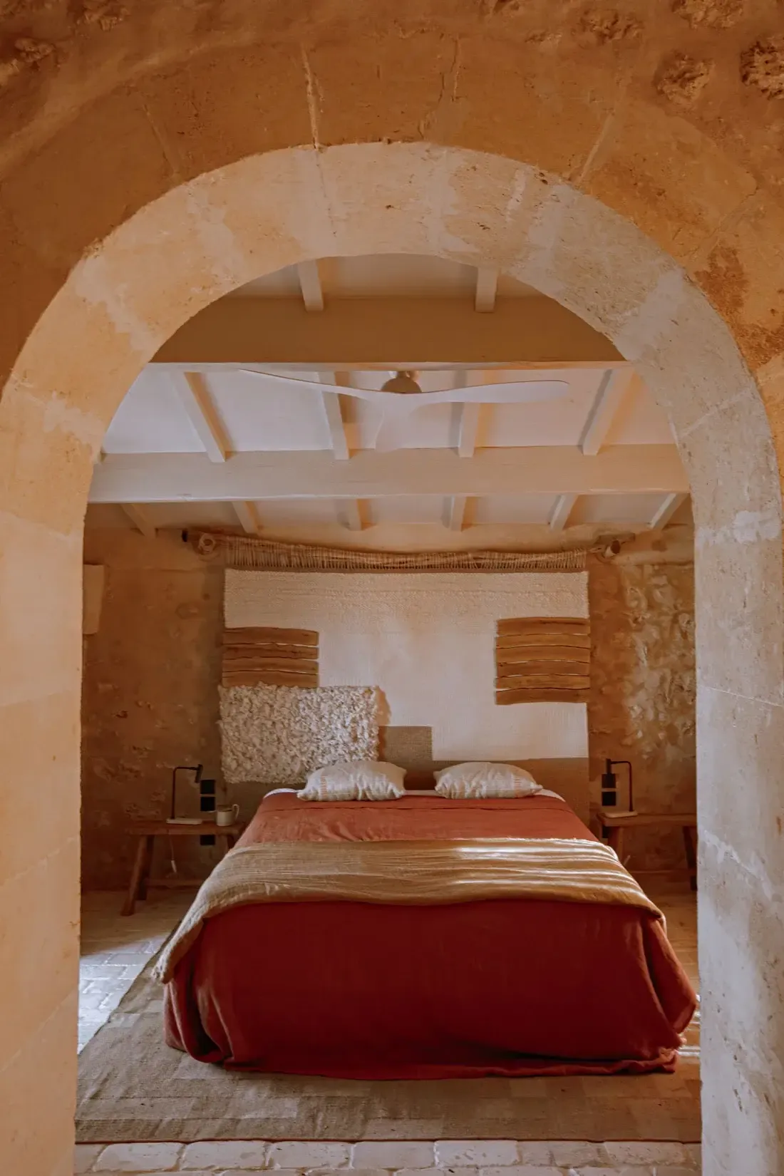 Son Blanc Farmhouse, Balearic Islands, Spain — Arched stone doorway framing bedroom with terracotta walls, wooden beams, red linen, and minimalist Mediterranean design.