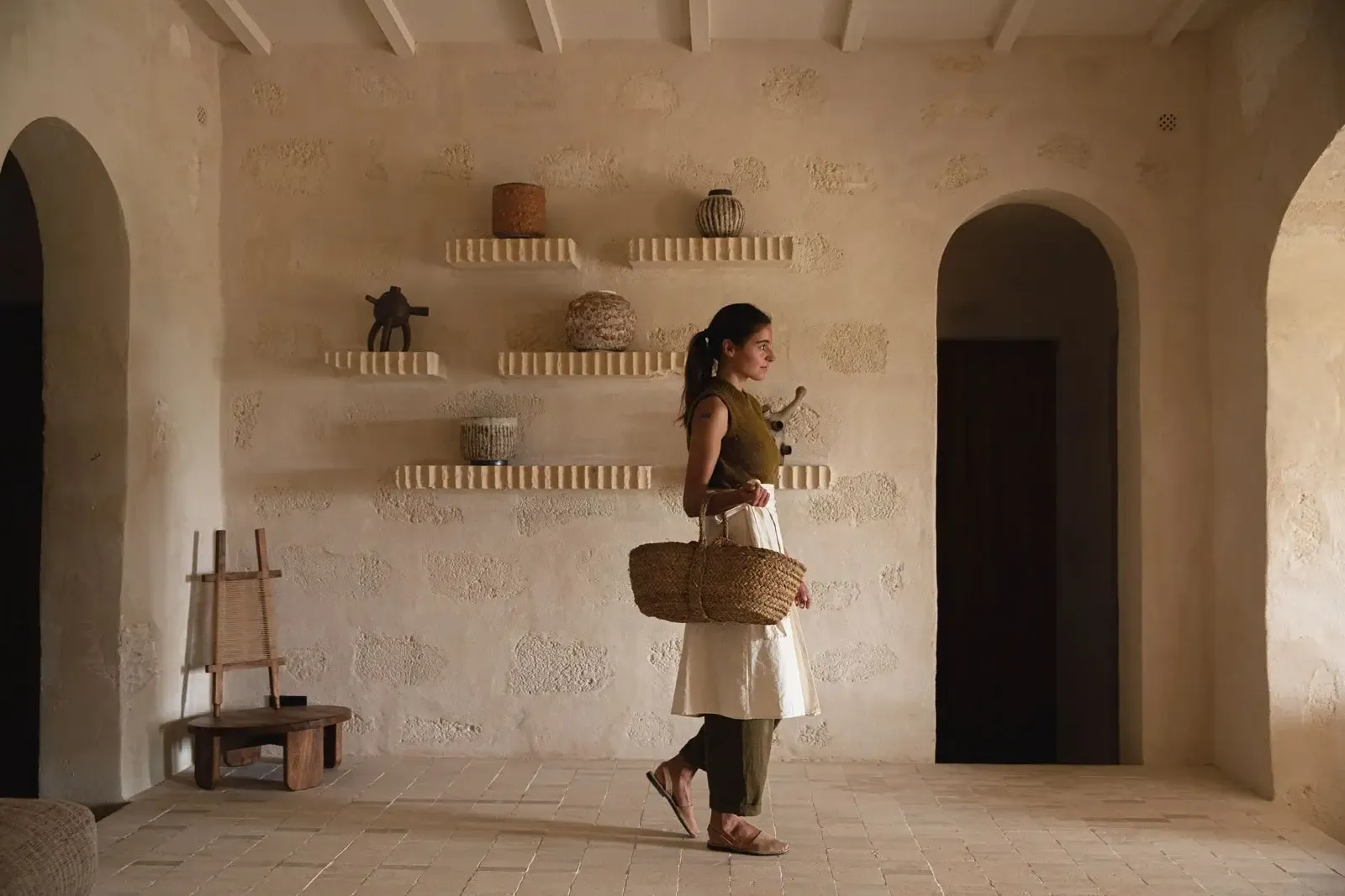 Son Blanc Farmhouse, Balearic Islands, Spain — Woman in linen holding woven basket in minimalist whitewashed room with arched doorways and floating shelves displaying ceramics.