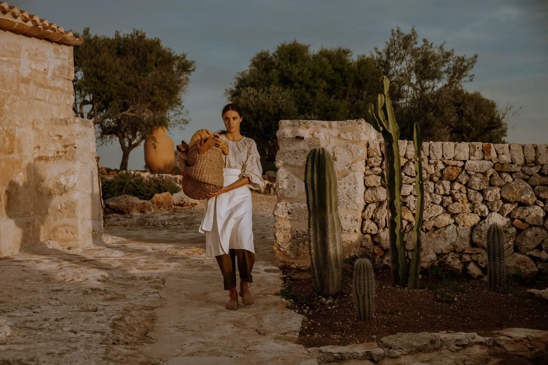 Son Blanc Farmhouse, Balearic Islands, Spain — Woman in linen dress holding woven basket in stone courtyard with cacti, terracotta vessels, olive trees, golden hour light.