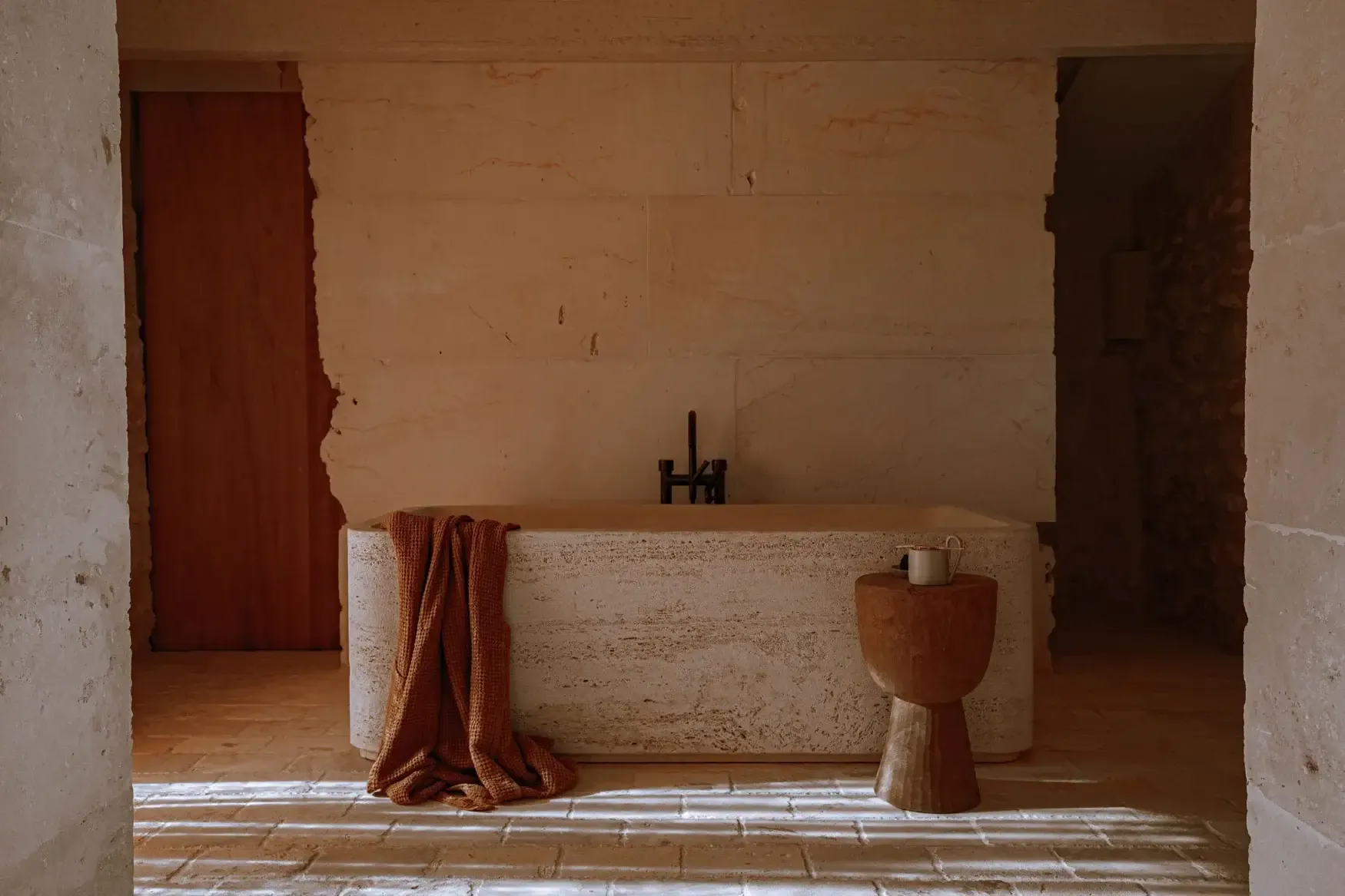 Son Blanc Farmhouse, Balearic Islands, Spain — Minimalist bathroom with travertine bathtub, wooden pedestal sink, terracotta textiles, and ochre-washed stone walls.