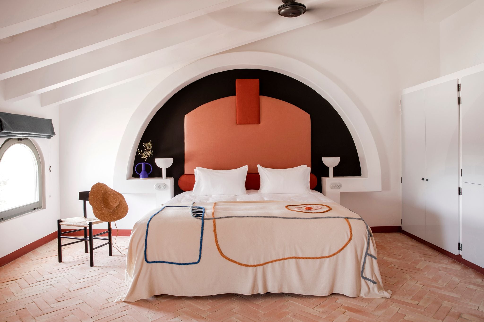 Menorca Experimental, Balearic Islands — Modern bedroom with terracotta headboard, arched niche, white linens, geometric throw blanket, wooden flooring, minimalist design.