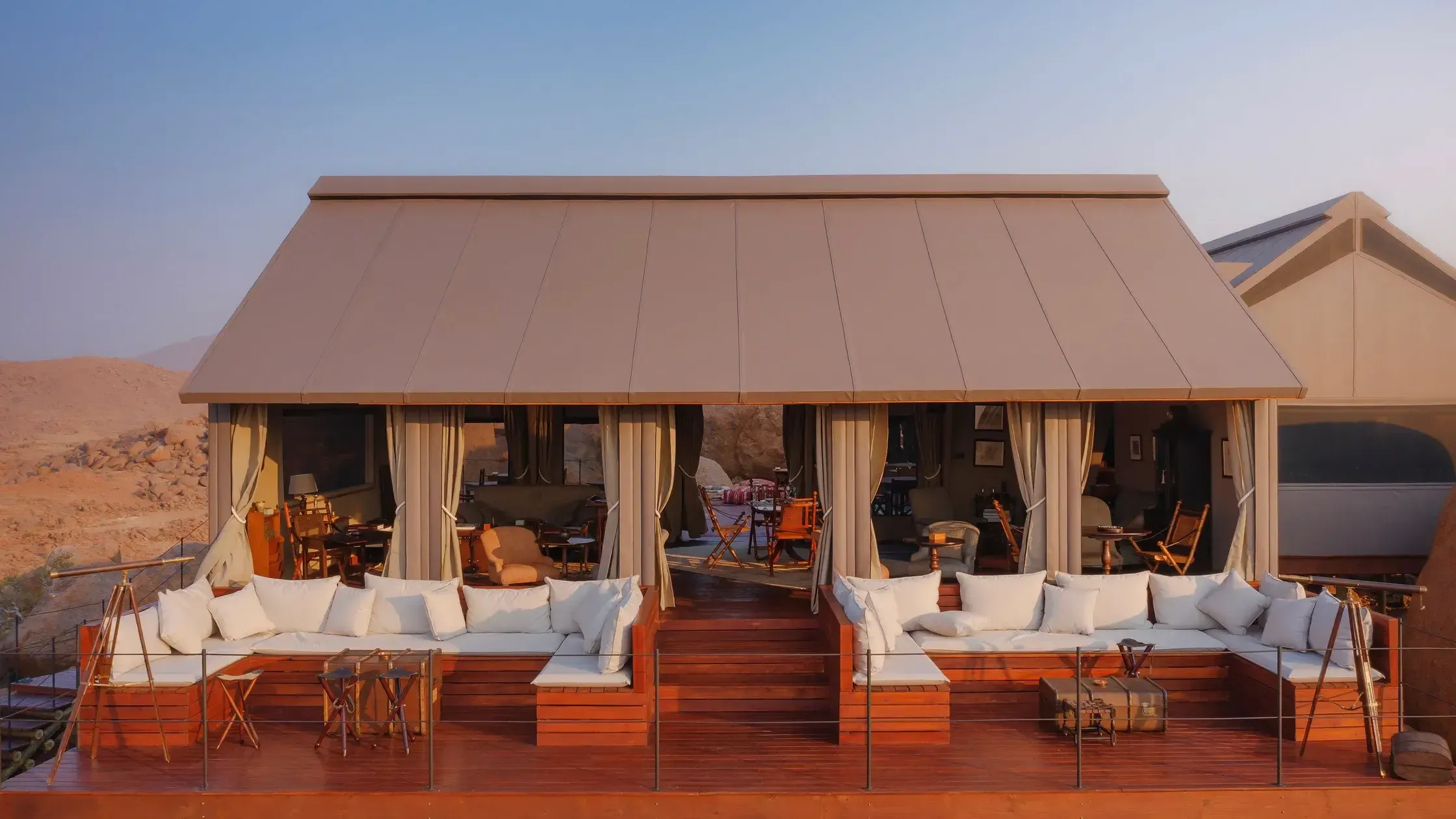 Zannier Sonop, Africa — Luxury safari lodge with taupe canvas awning, wooden deck, cream cushioned seating, floor-to-ceiling curtains, and desert landscape at sunset.