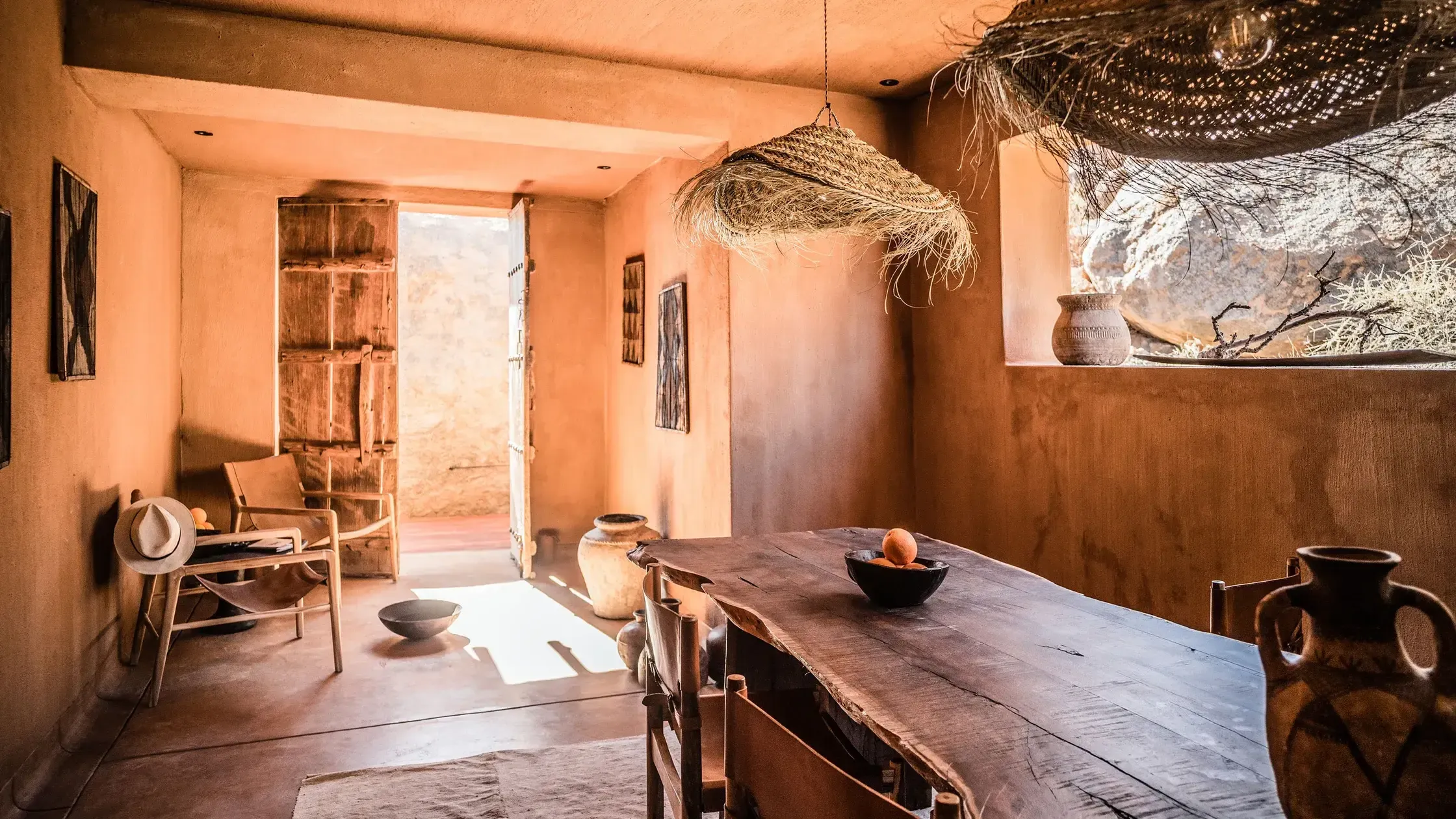 Zannier Sonop, Africa — Ochre-walled dining room with wooden table, woven pendant lights, ceramic vessels, and desert landscape views through open doorway.