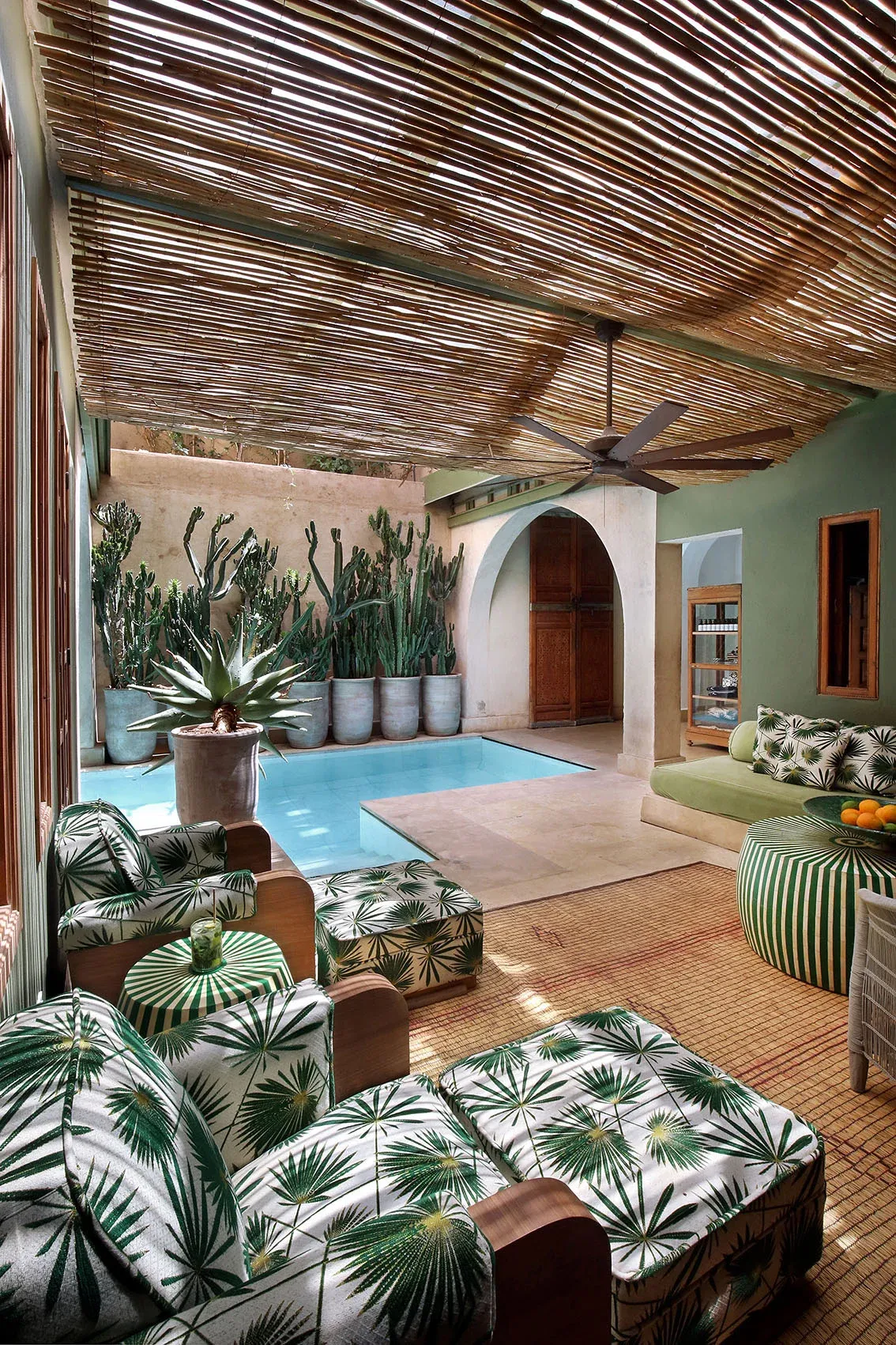 El Fenn, Marrakech, Morocco — Courtyard with blue plunge pool, palm-patterned furnishings, slatted timber ceiling, cacti in ceramic pots, arched doorway, and ceiling fan.