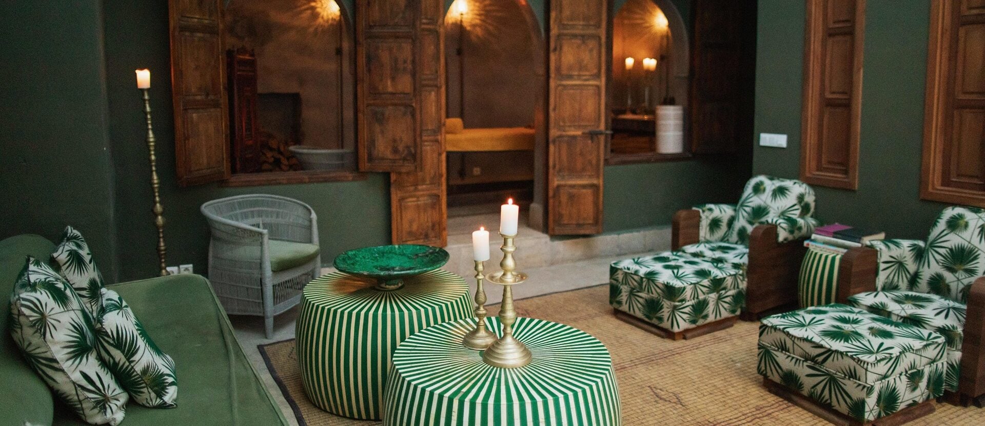 El Fenn, Marrakech, Morocco — Green striped ottomans with palm-print pillows, brass candelabra, arched wooden doors, candlelit courtyard lounge.