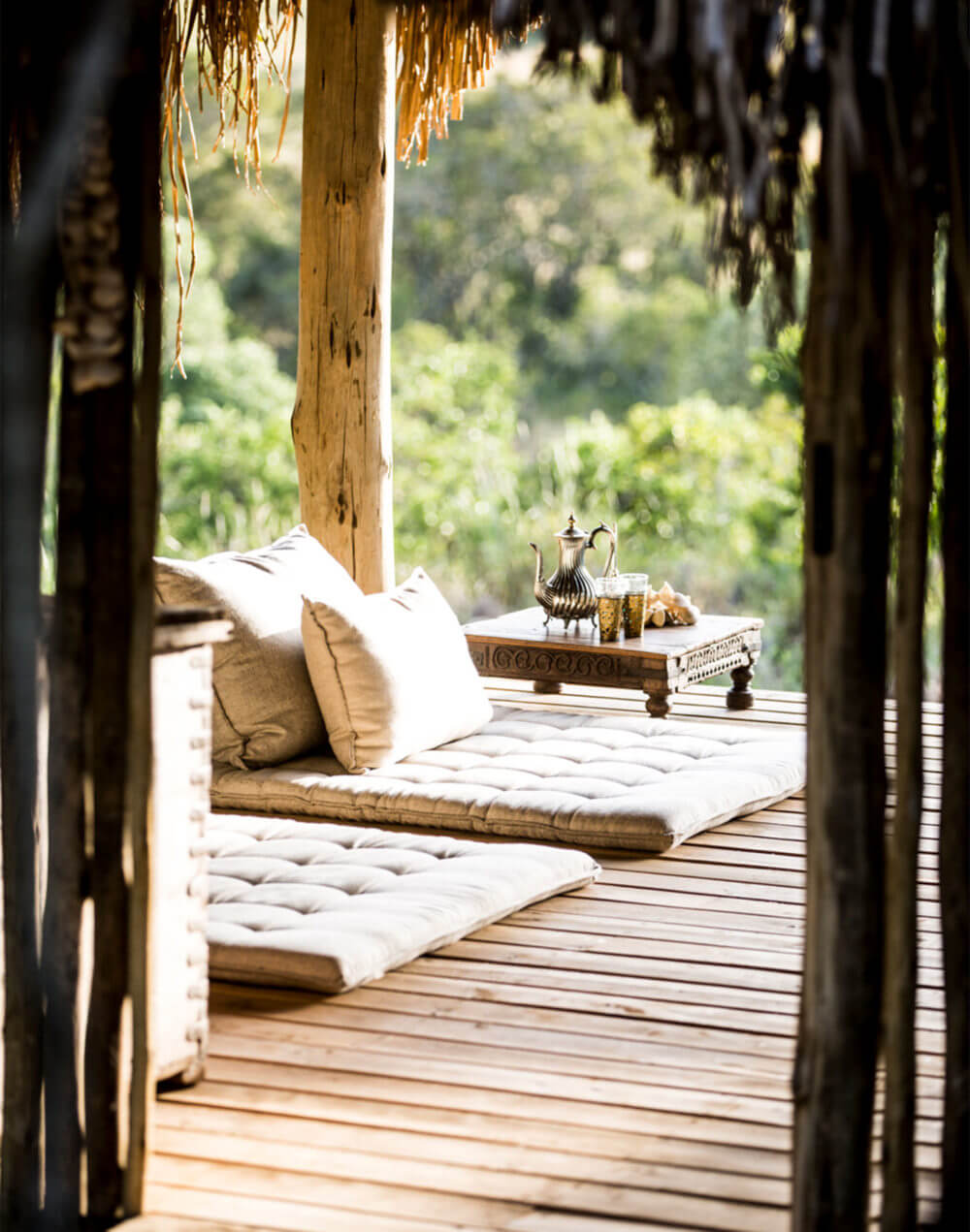 Spa retreat with wooden deck, white cushioned daybed, brass teapot, and mountain vista through open doors, natural light and tropical forest backdrop.